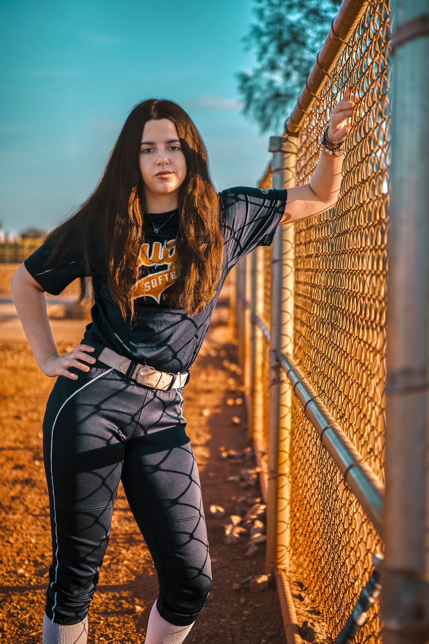 Young female softball player in uniform leaning against chain-link fence at baseball field during golden hour.
