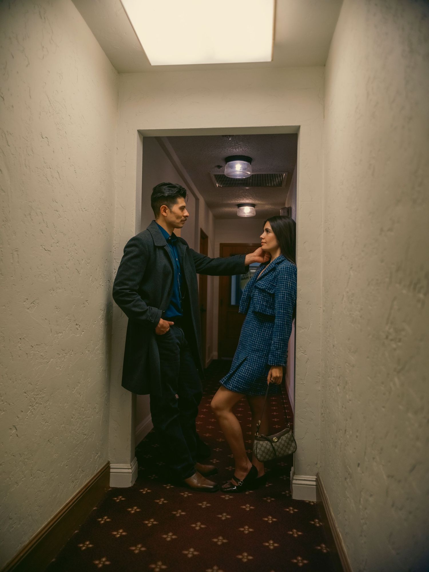 A couple in blue outfits stands in a hotel hallway, the woman resting her hand on the man's chest.