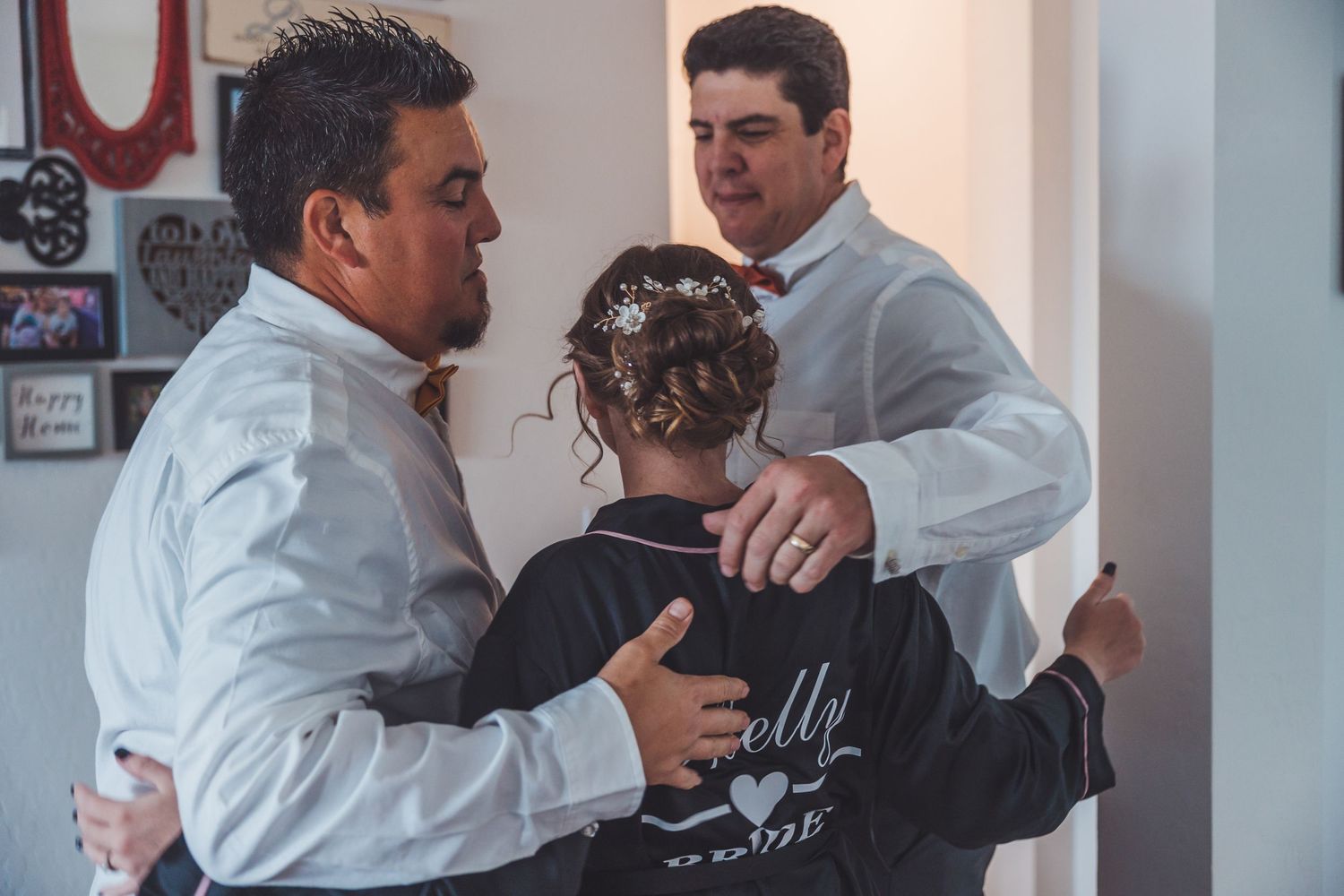 Two men in white dress shirts help a bride with her wedding jacket in a warmly decorated home.