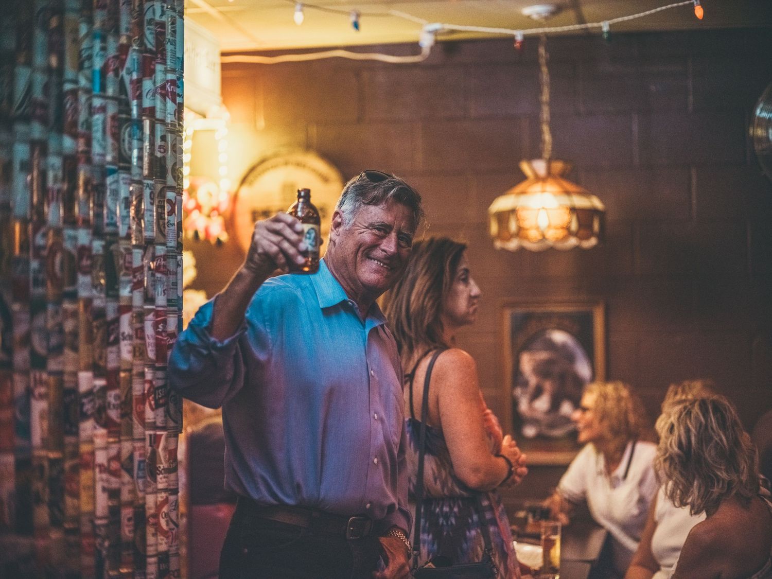 Man smiling and holding a bottle at a lively bar gathering with friends in a warmly lit venue.