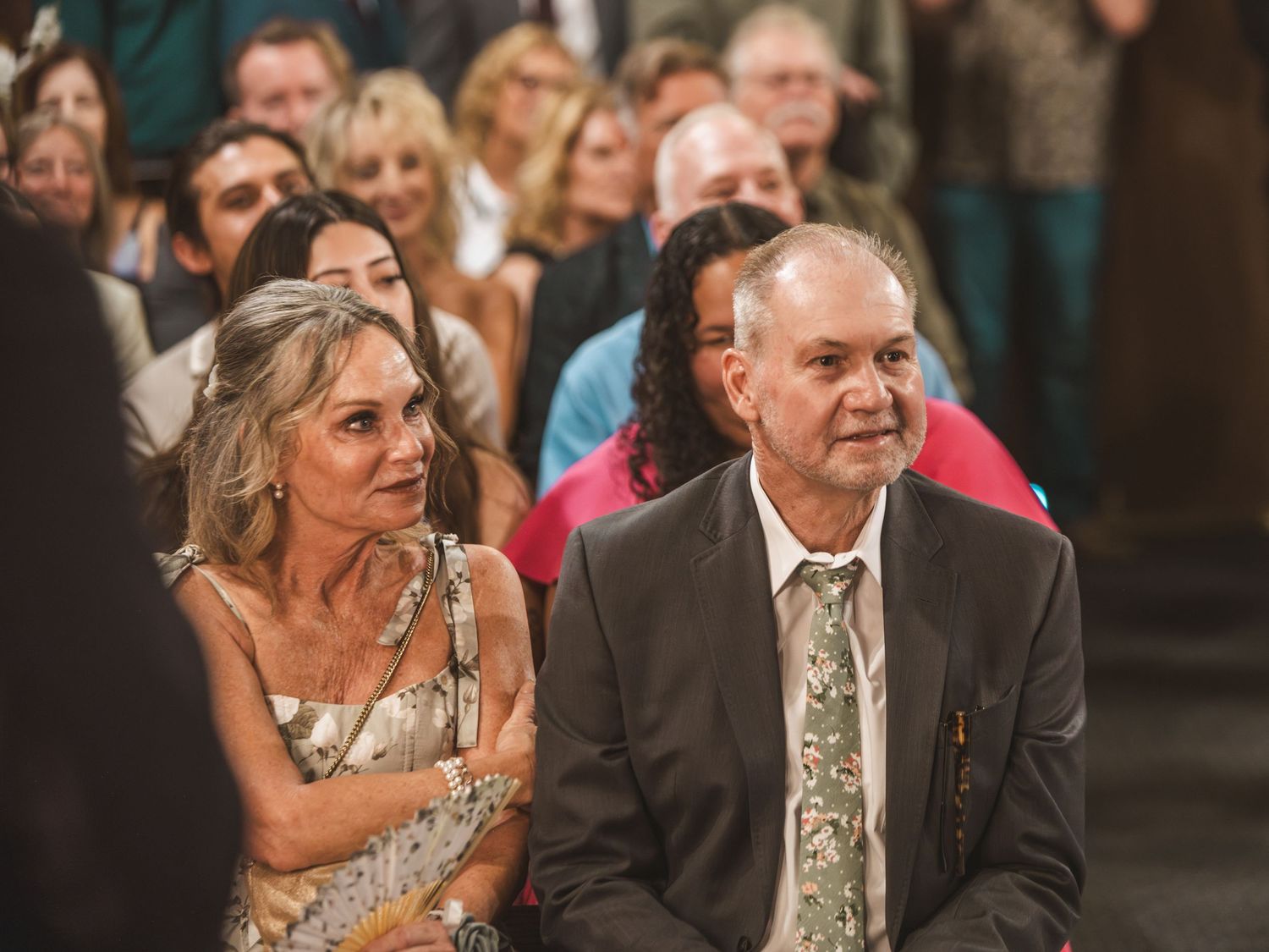 Couple seated at a wedding ceremony, woman in floral dress and man in suit with tie, guests in background.