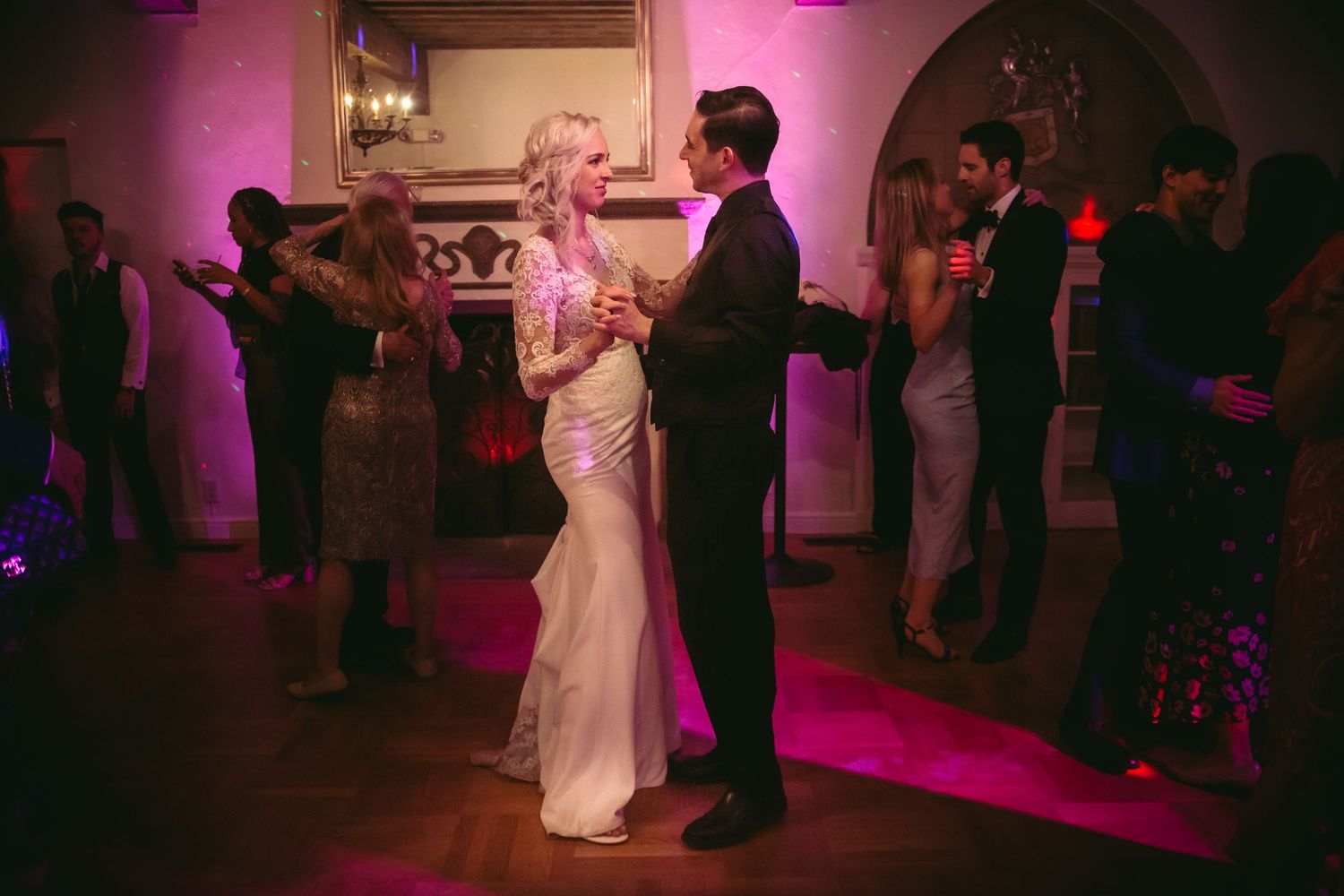 Bride in white lace gown and groom dancing together in a pink-lit elegant wedding reception hall.