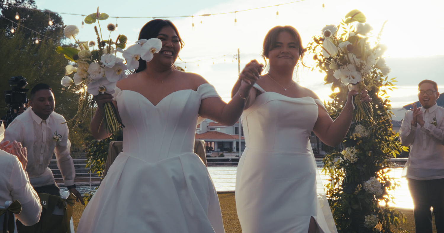 Two brides in white gowns walking together at golden hour celebration.