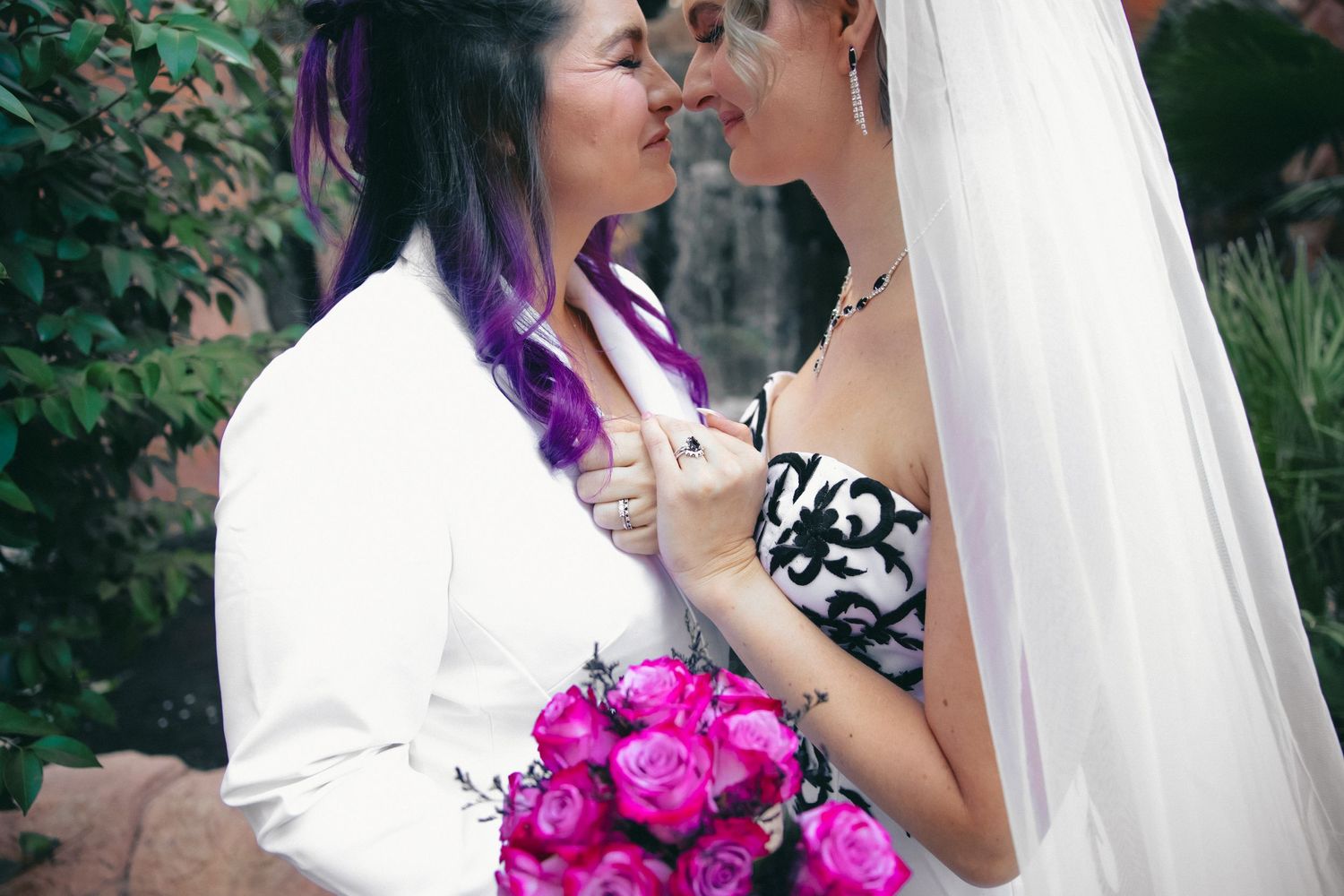 Two brides nearly kiss while holding vibrant pink roses bouquet in a garden setting.