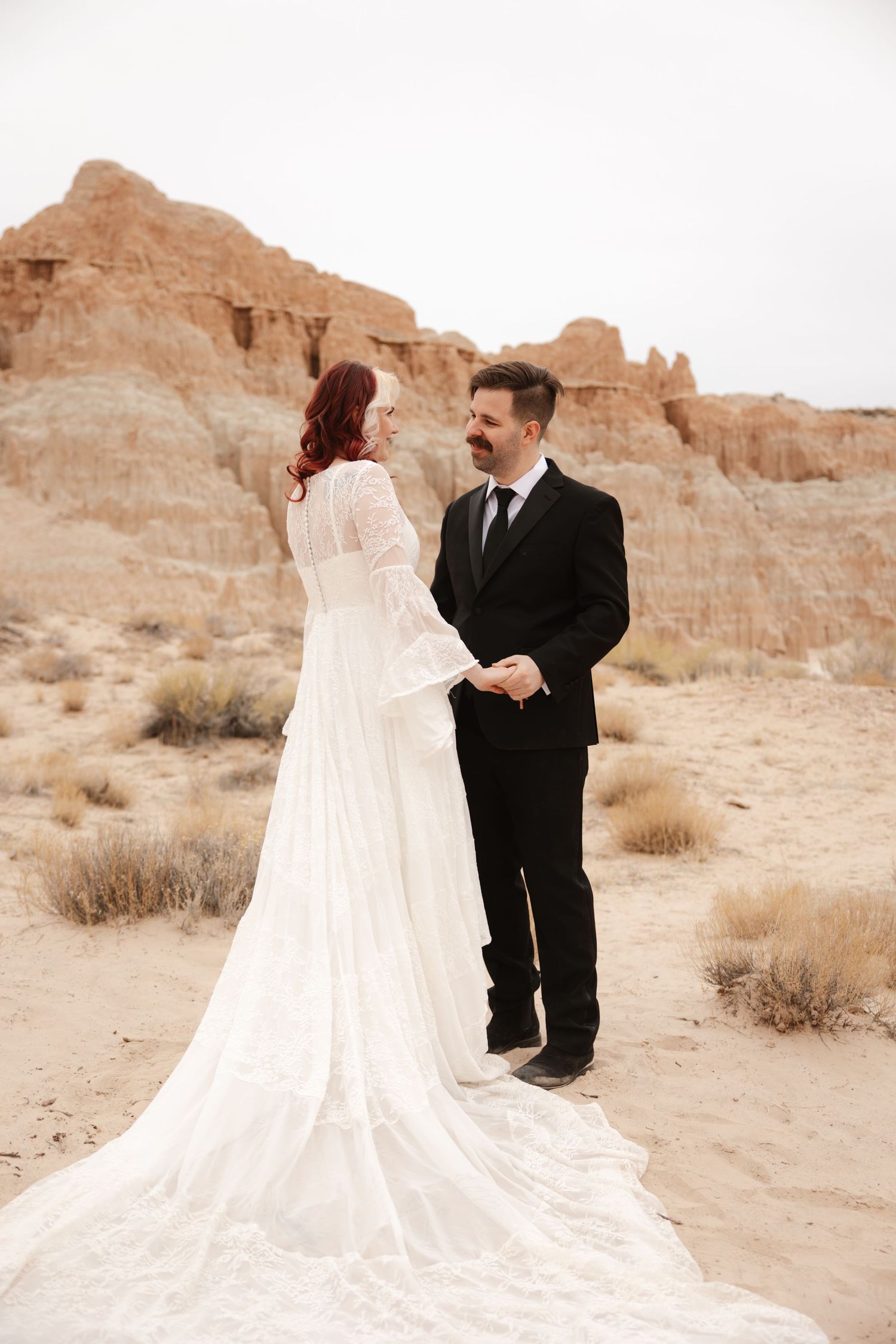 Bride and groom hold hands facing each other in sandy desert with eroded rock formations behind them.