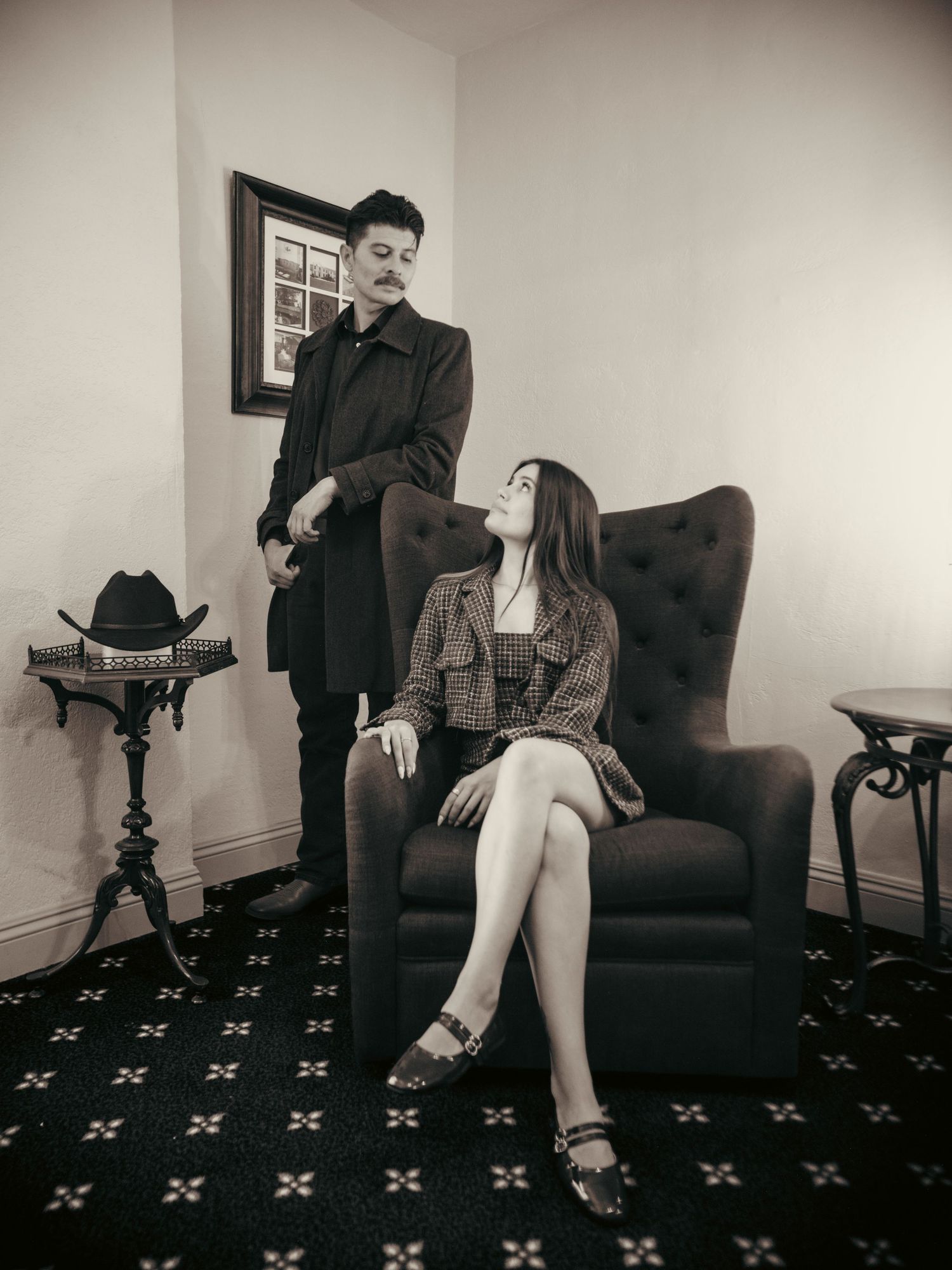 Black and white photo of a man standing beside a woman seated in a velvet armchair in an elegant room.