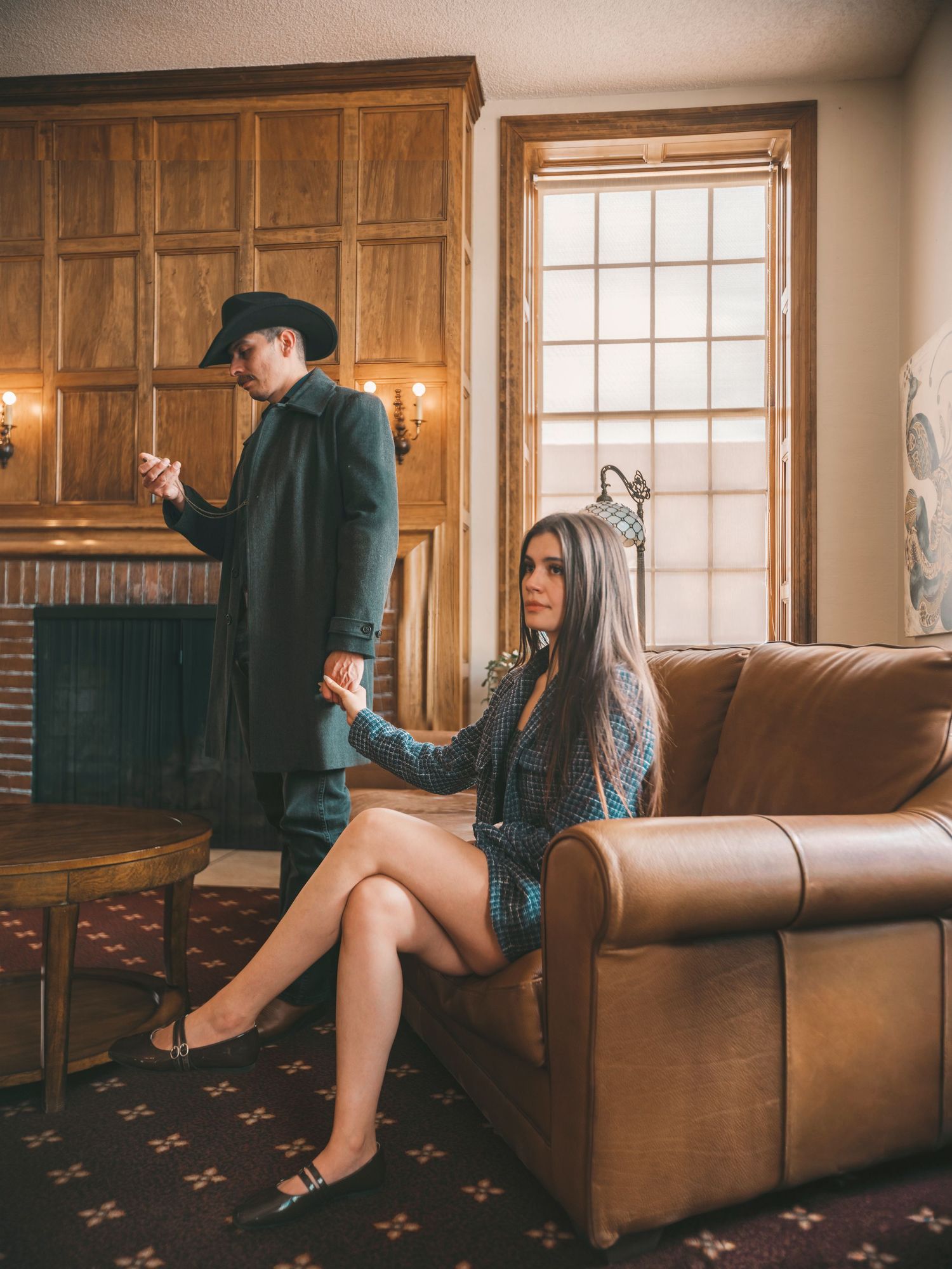 A woman sits on a leather sofa while a man in a cowboy hat stands by a fireplace in a rustic wood-paneled room.