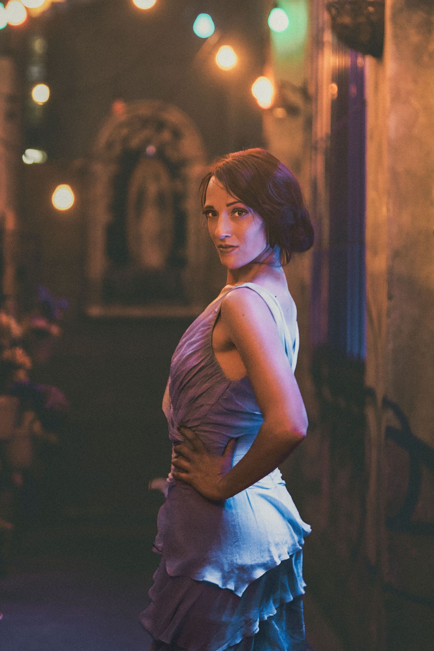 Woman in elegant dress poses confidently in dimly lit venue with warm string lights in background.