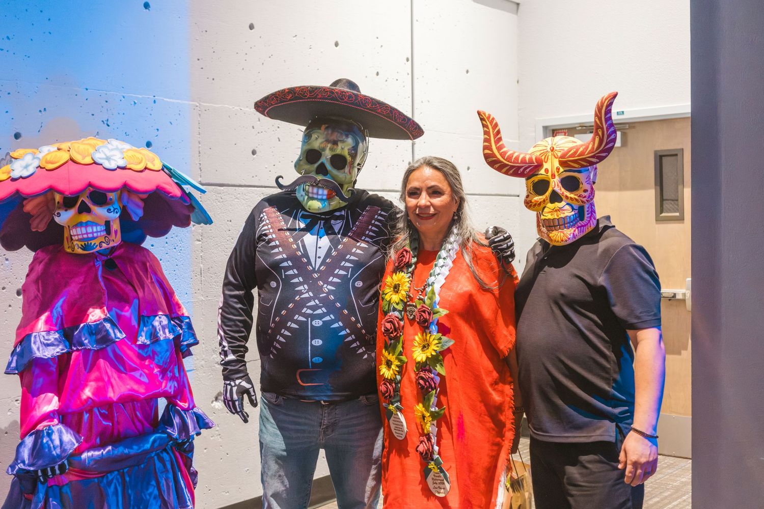 Woman in orange dress poses with two people in colorful Day of the Dead skull masks and costumes at festive event.
