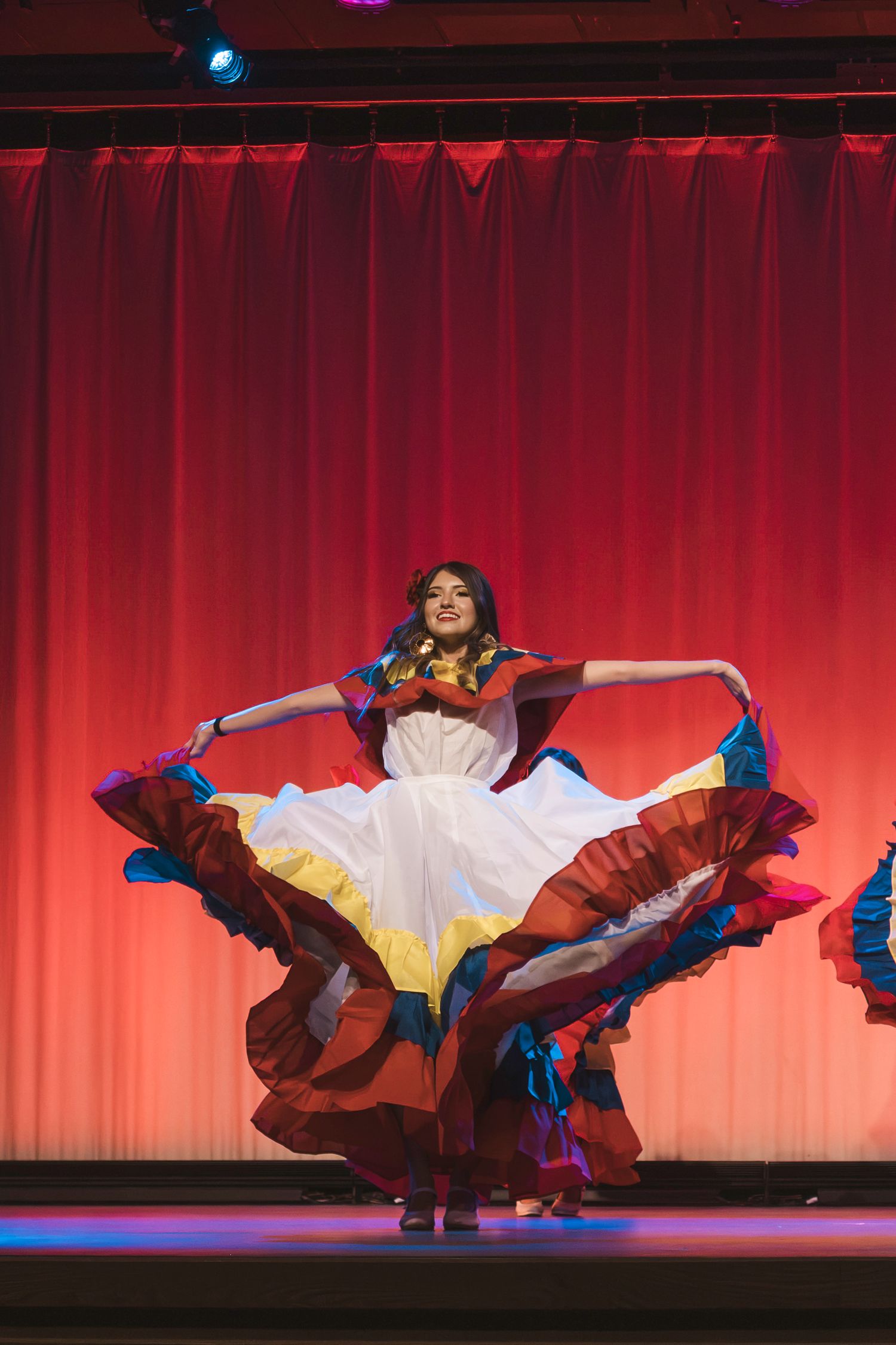Dancer in colorful traditional folk costume twirling on stage with red curtain backdrop.