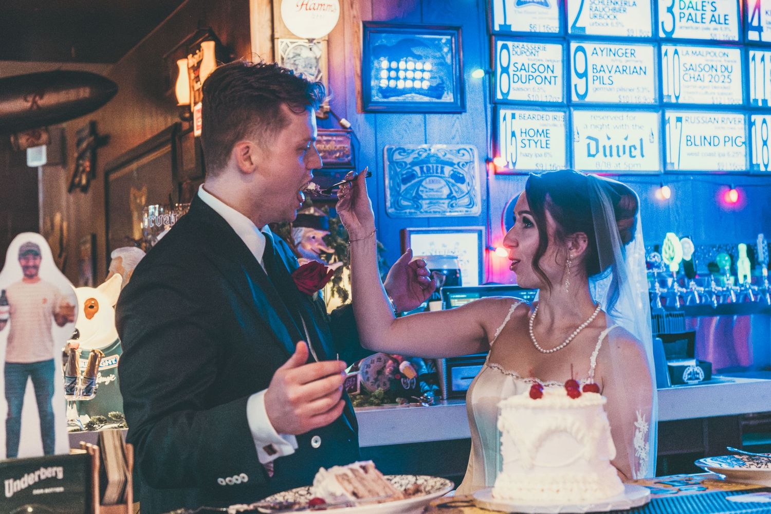 Bride and groom toasting with drinks beside a wedding cake in a bar decorated with neon signs.