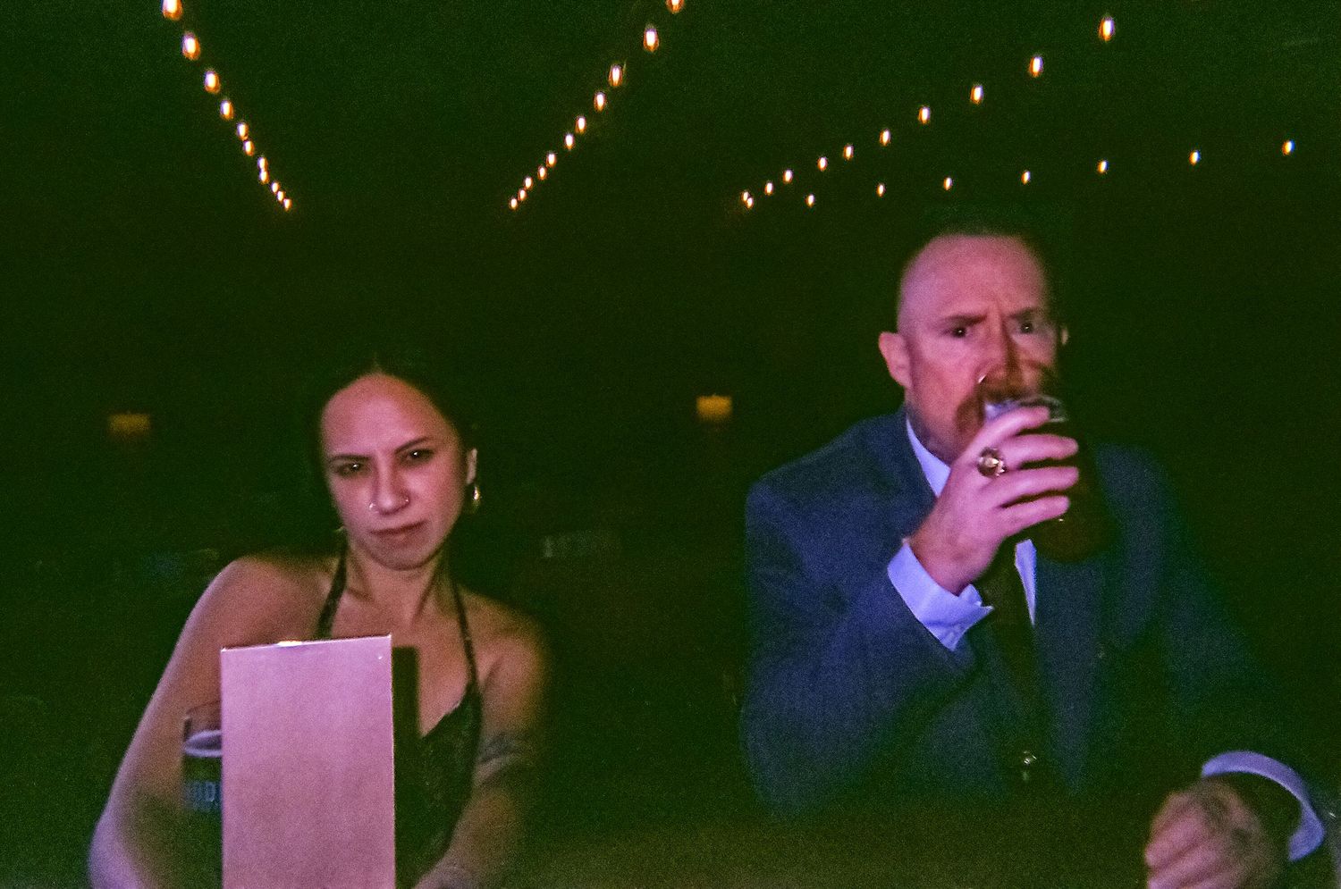 A man in a suit drinking and a woman in a strapless dress sit together at an outdoor evening event.