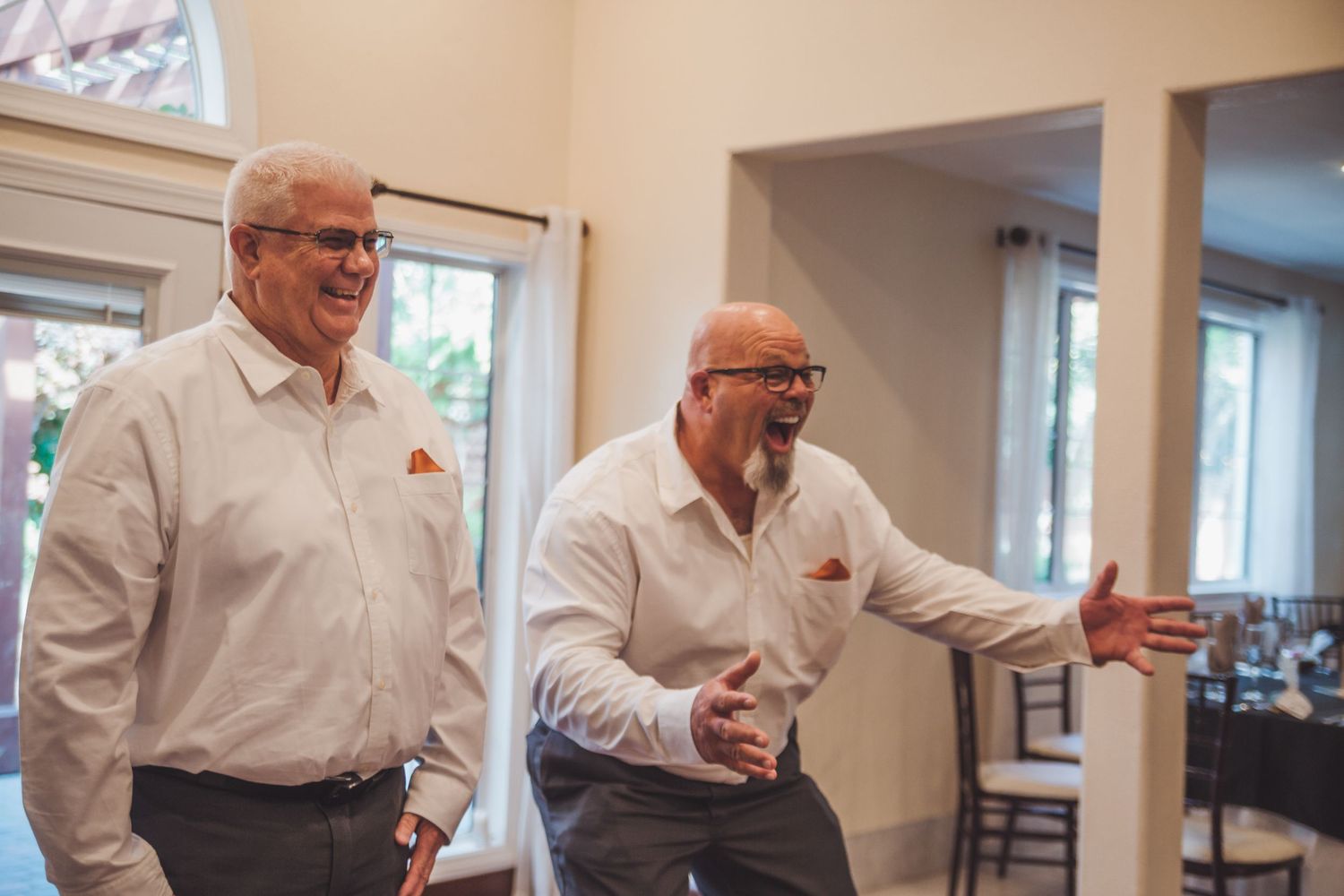 Two men in white dress shirts with orange pocket squares laugh together inside an elegant wedding venue.