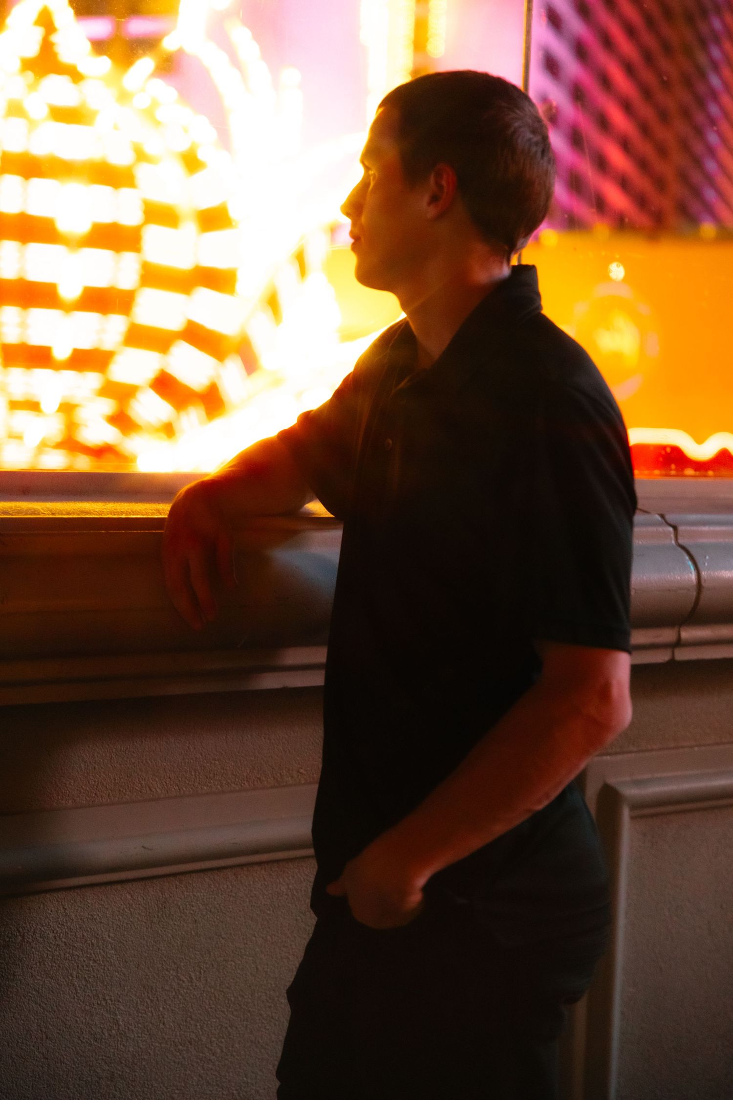 Man in black polo shirt leans against wall bathed in warm golden neon light at night in Las Vegas.