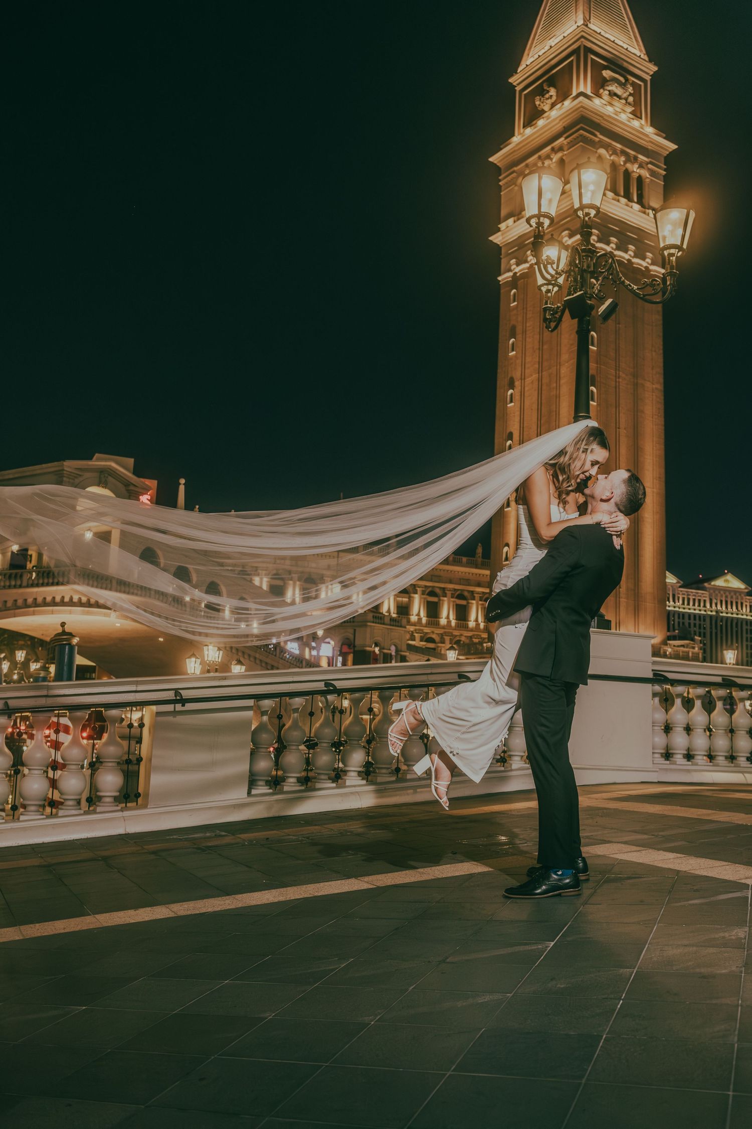 Groom sweeping bride off her feet with veil flowing beside The Venetian Las Vegas at night.