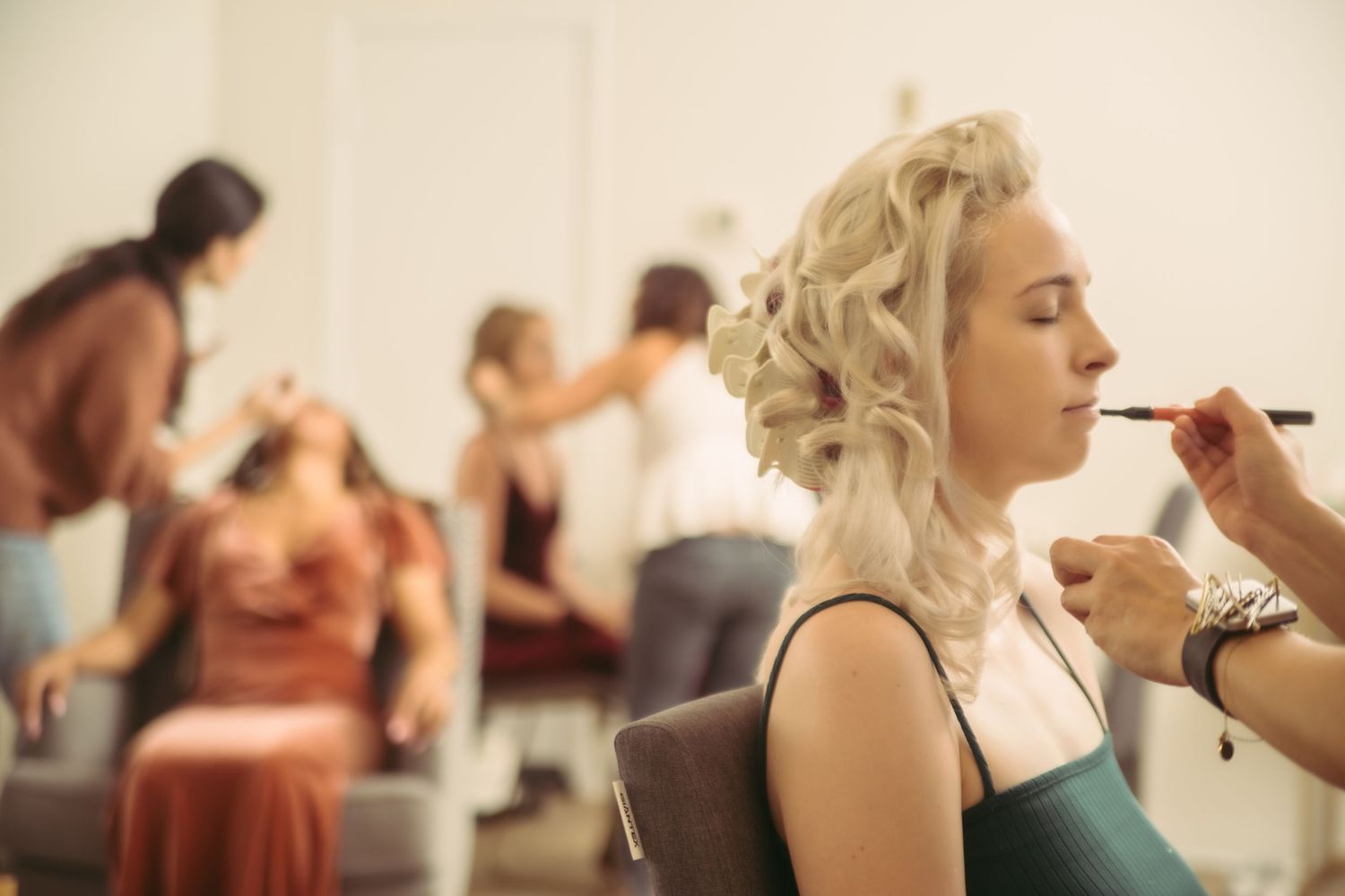 A makeup artist applies lipstick to a blonde woman with curly hair in a busy salon mirror setting.