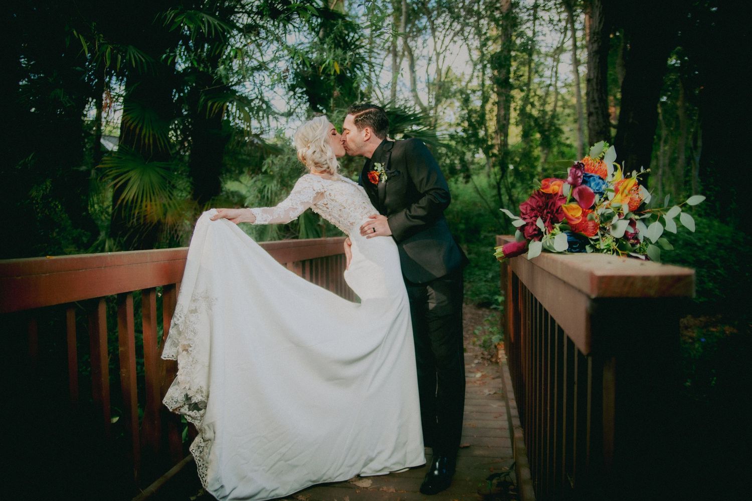 Bride and groom sharing a romantic kiss on a wooden bridge surrounded by lush green trees, with a colorful bouquet nearby.
