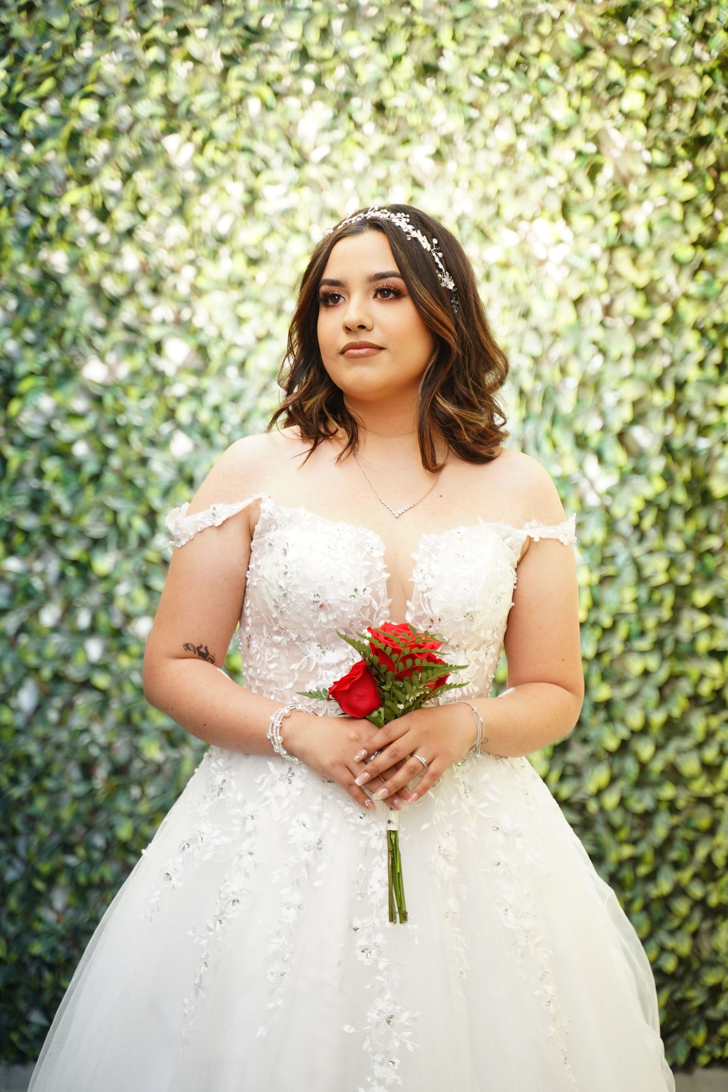 Bride in white lace off-shoulder ball gown holding red roses bouquet, posing before green leaf wall backdrop.
