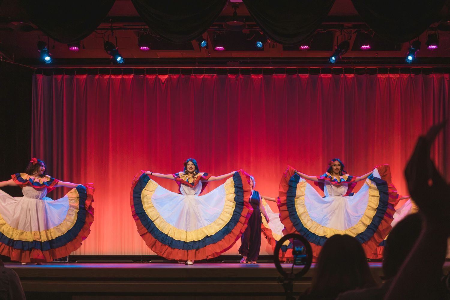 Folkloric dancers in colorful traditional dresses perform on a red-curtained stage during a vibrant cultural show.