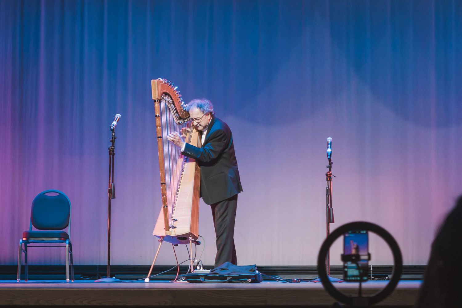 Musician playing a wooden harp on stage during a live concert performance with microphones and blue stage lighting.