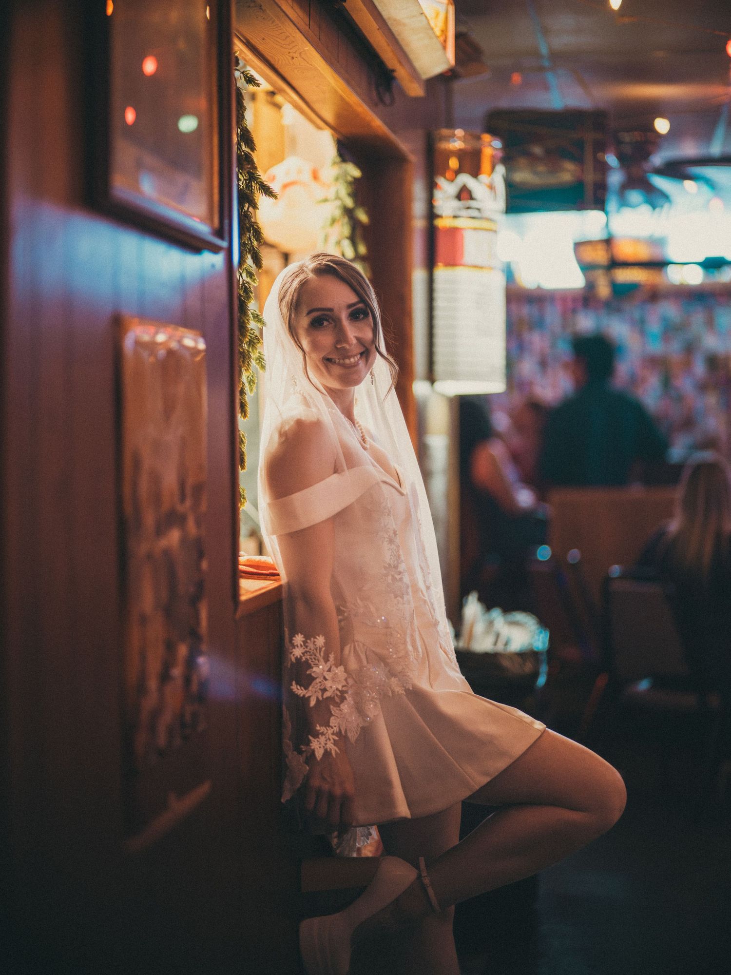 Bride in short wedding dress and veil posing in a warmly lit bar, smiling over her shoulder.