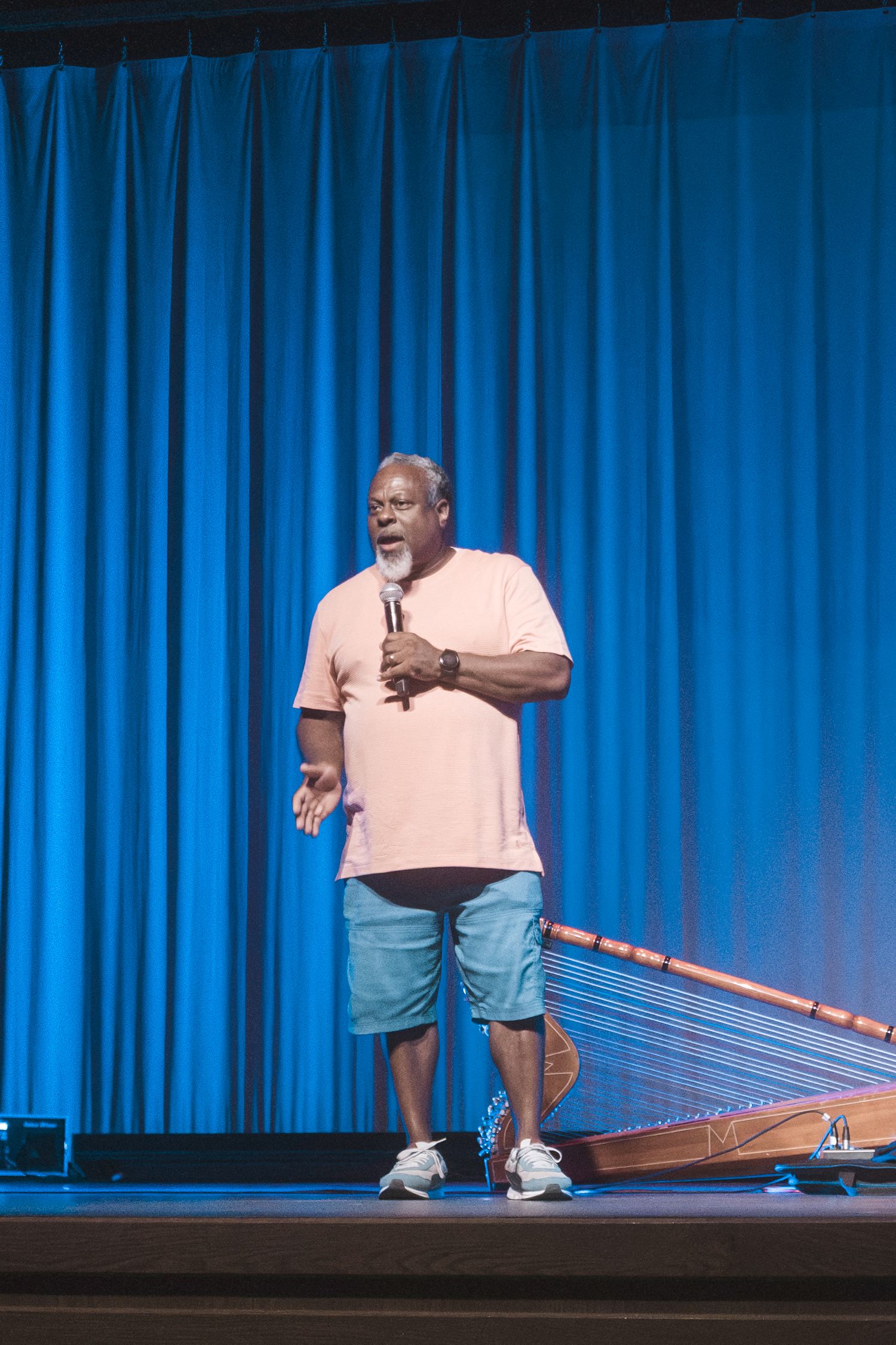 Man in pink shirt and blue shorts speaks into microphone on stage with blue curtain backdrop.