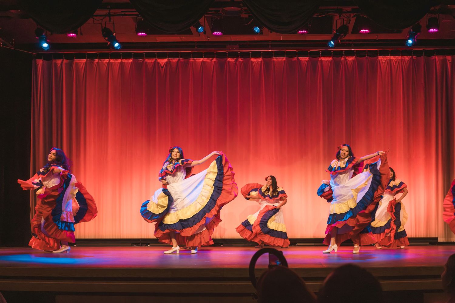 Colorful folkloric dancers perform on a red-curtained stage in vibrant traditional dresses.