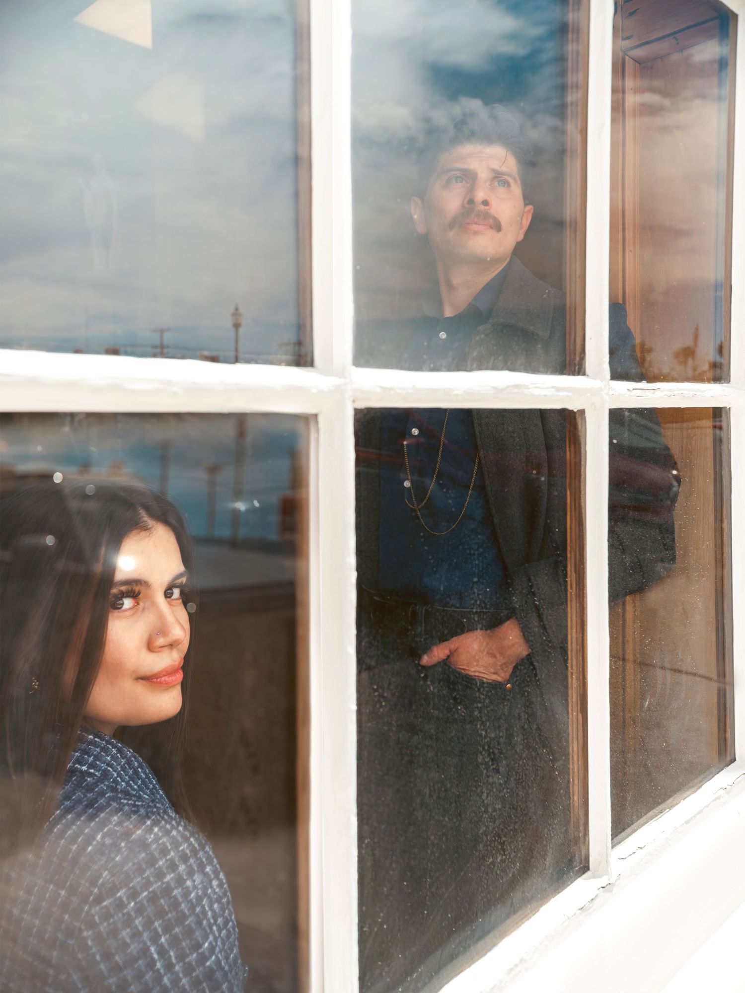 A man and woman gazing through a vintage white-framed window, reflecting sky and surroundings.