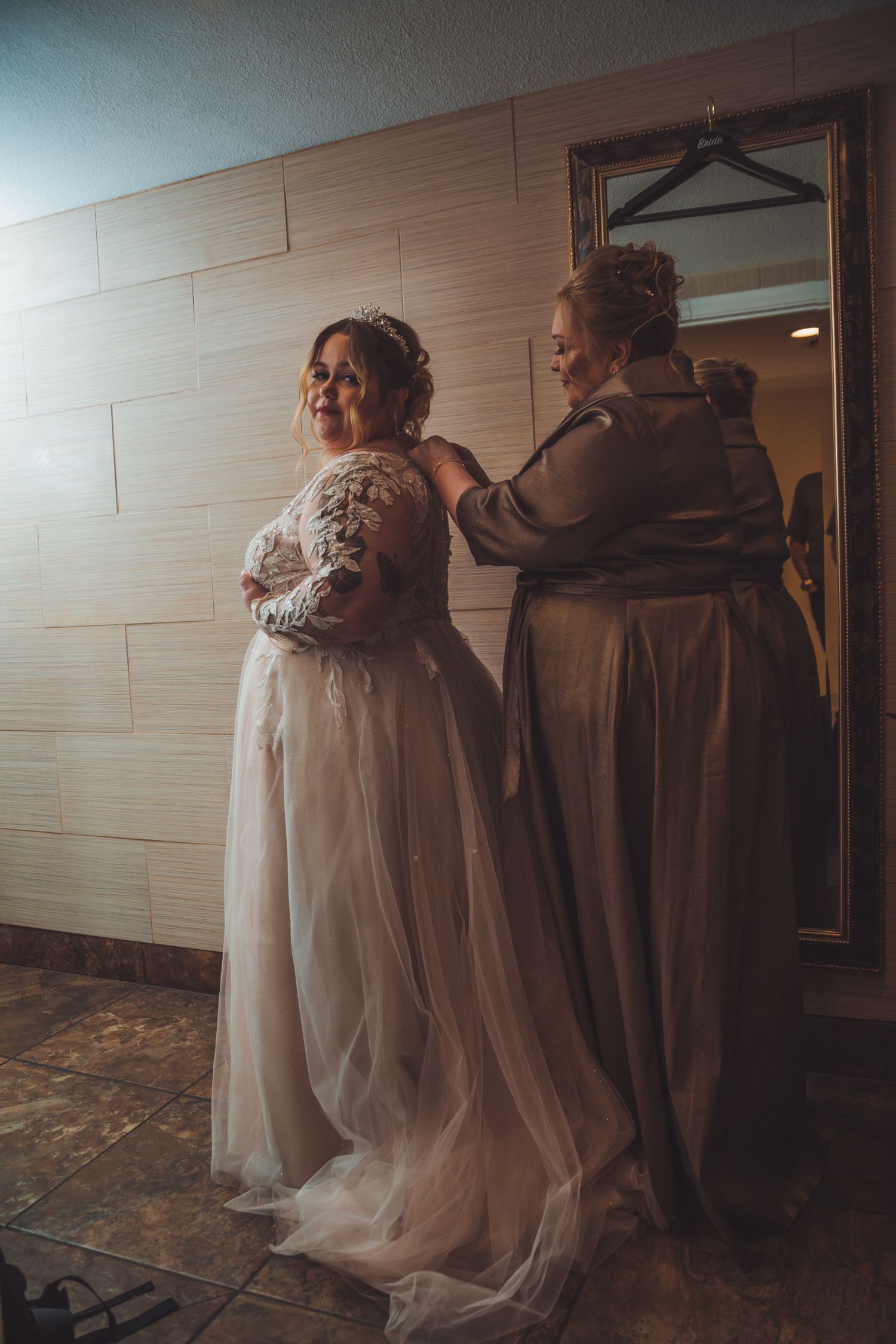 A bride in a lace wedding gown being helped into her dress by a woman near a gold mirror.