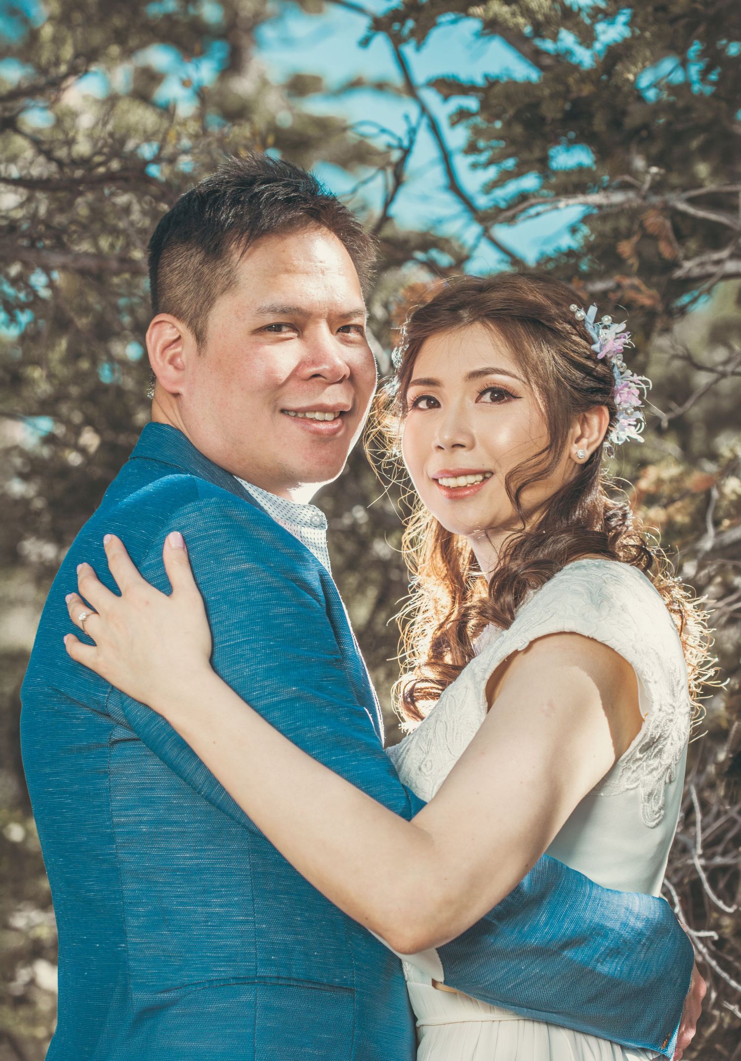 Smiling bride and groom hugging outdoors with pine trees background during sunny wedding photo session.