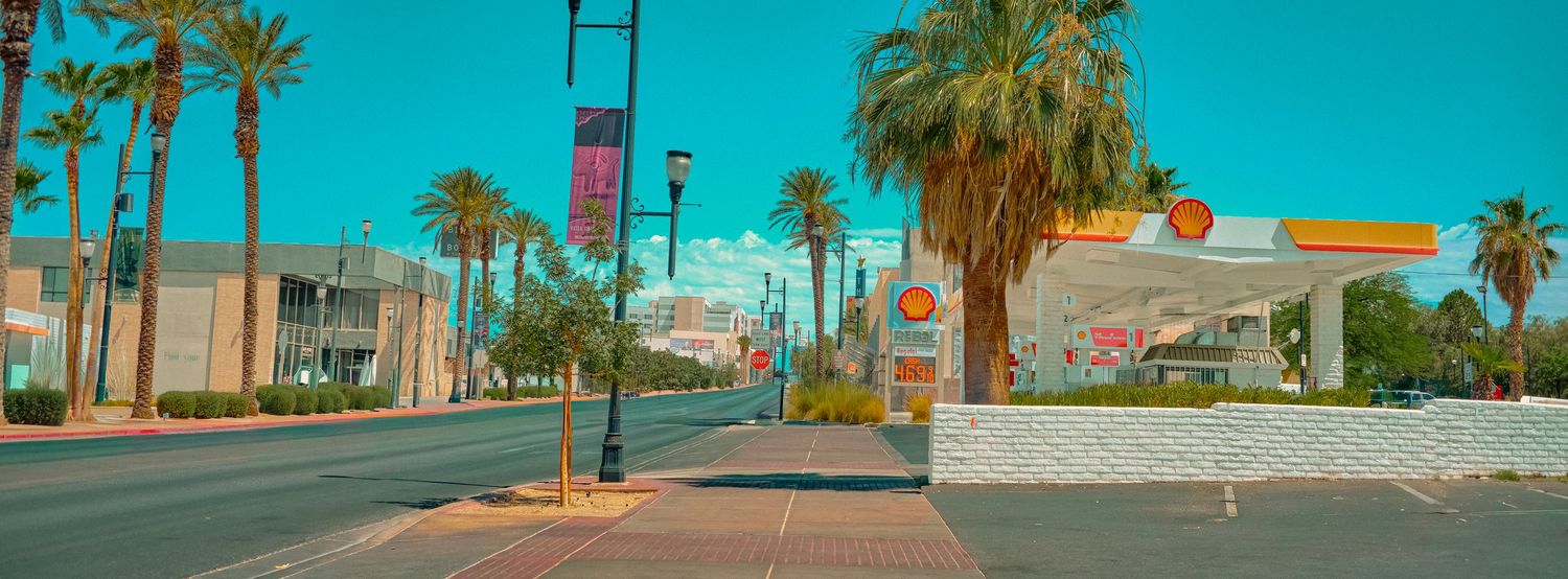 Sun-drenched street with palm trees, Shell gas station, and colorful flags under a vivid turquoise sky in a desert city.