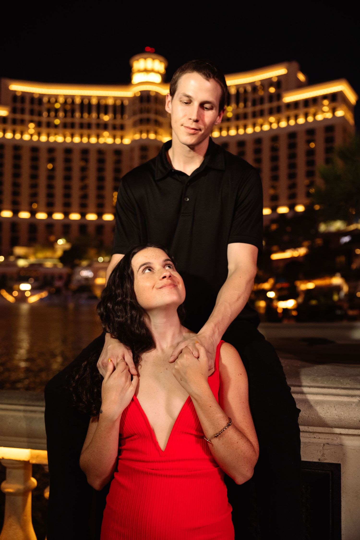 Woman in red dress looks up at man in black shirt posing together at night near Bellagio Las Vegas.