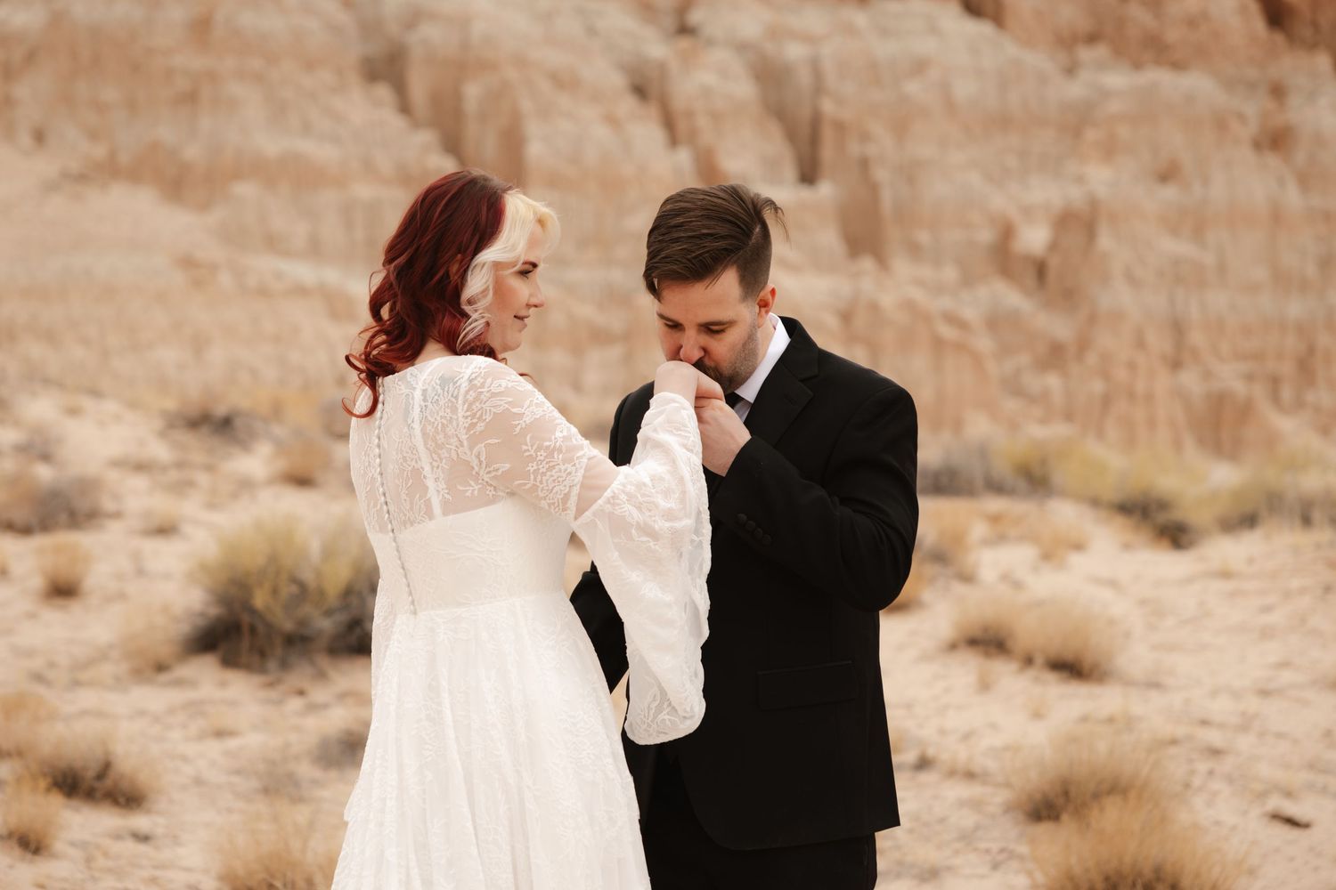 Groom kissing bride's hand during desert elopement ceremony with sandy rock formations in background.