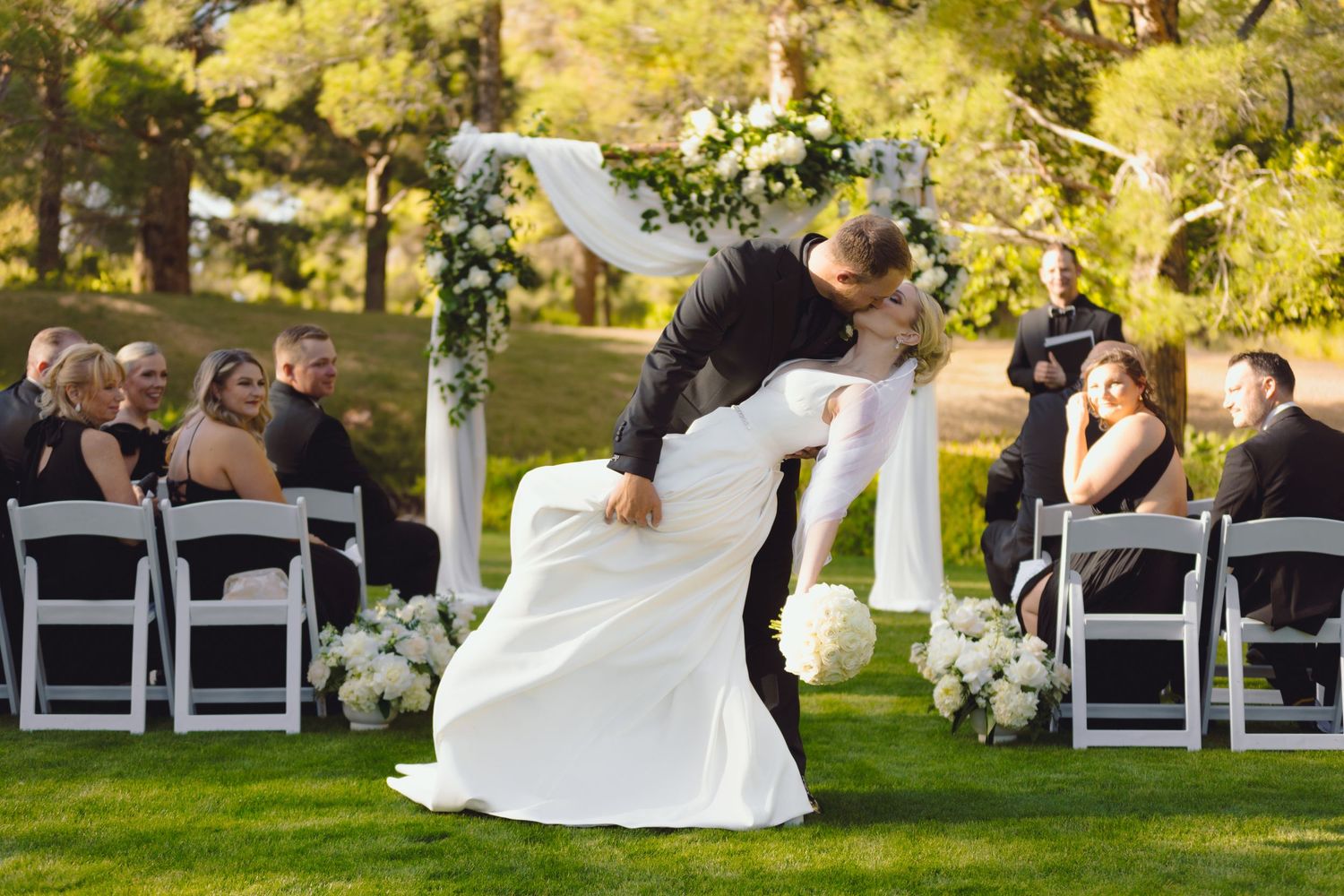 Groom dips bride for a kiss at outdoor wedding ceremony on green lawn.