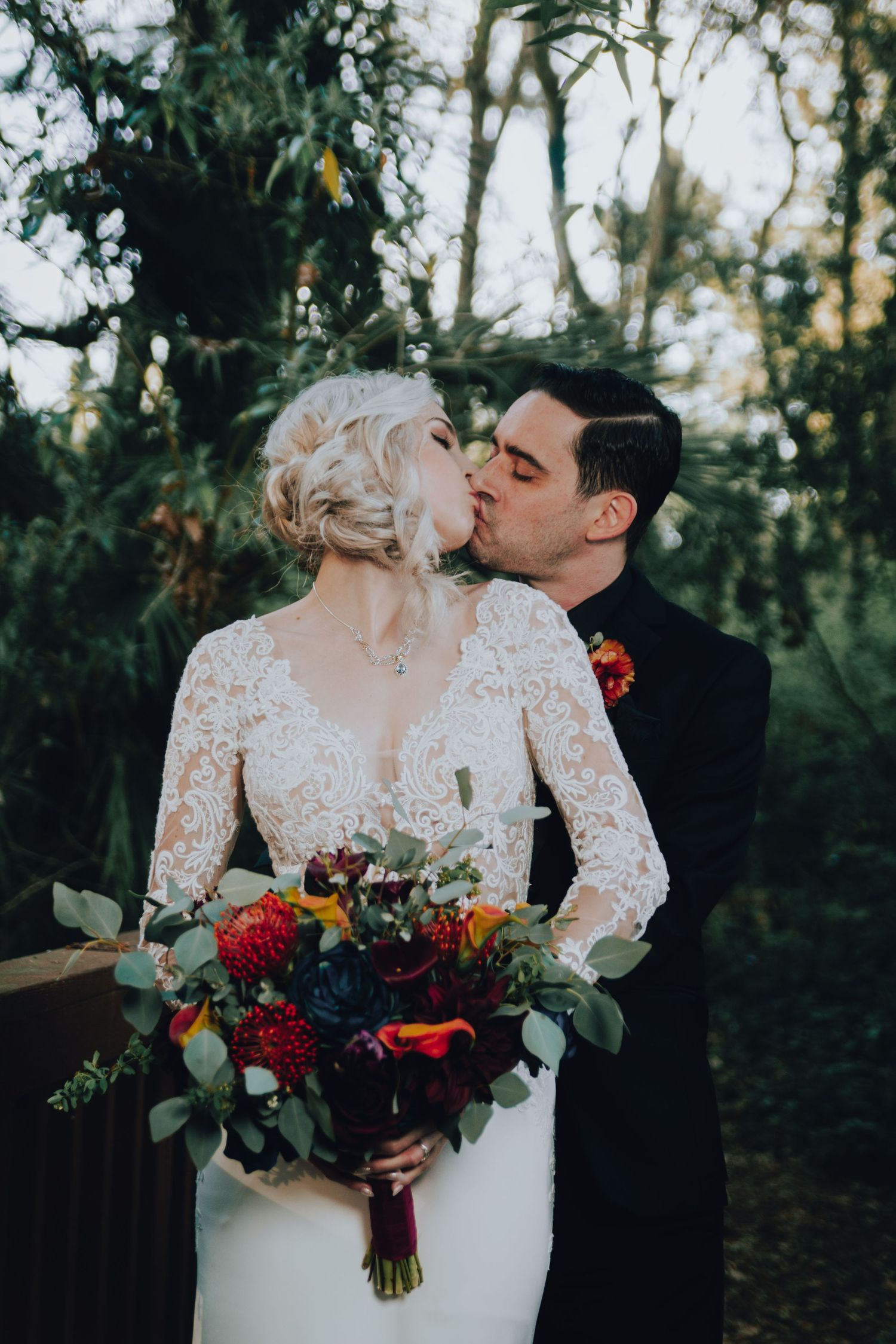 Bride and groom sharing a romantic kiss, bride holding a vibrant bouquet of red, orange, and purple flowers with eucalyptus.