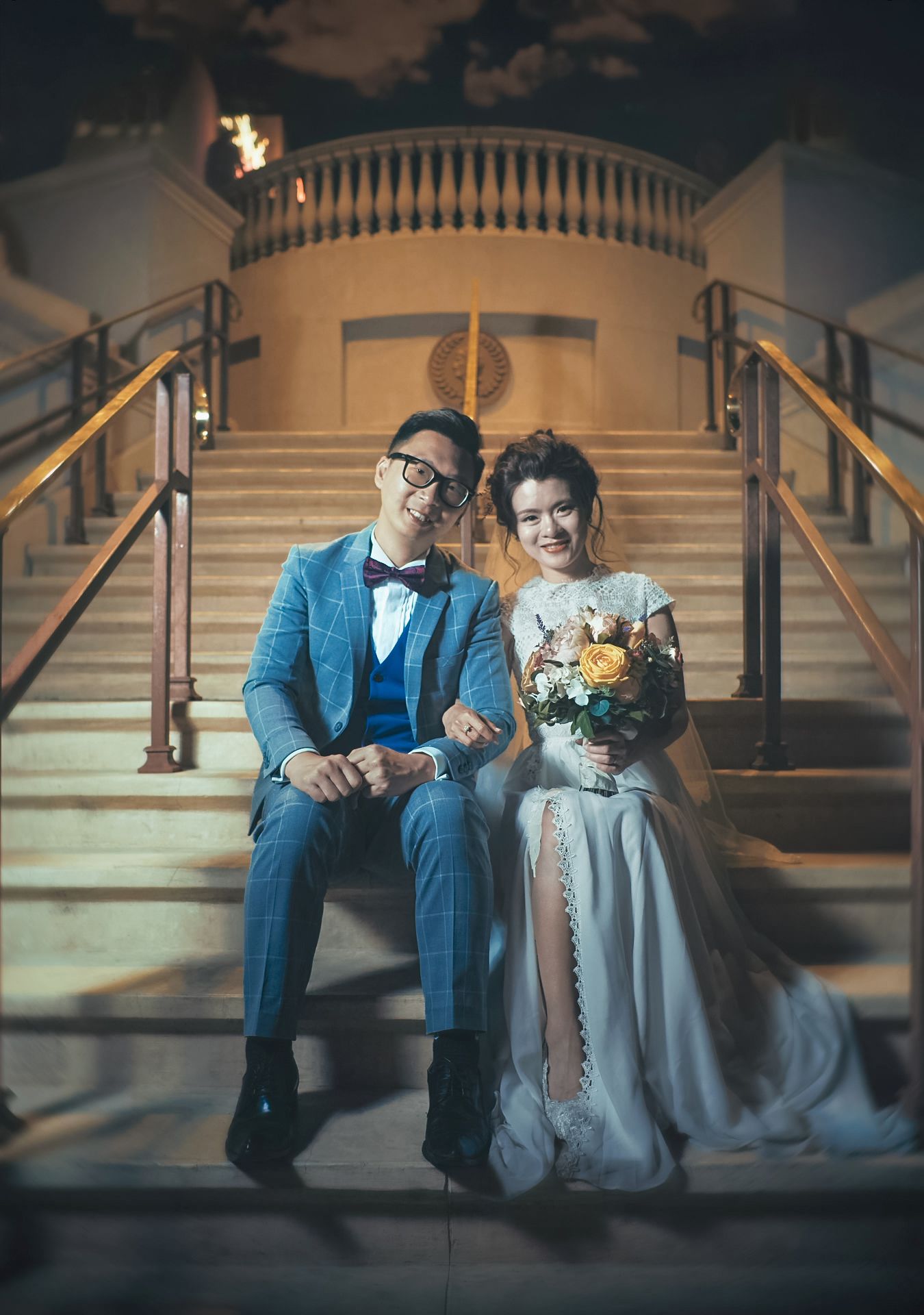 Couple in wedding attire sitting on grand staircase, groom in blue suit, bride in white gown holding bouquet.