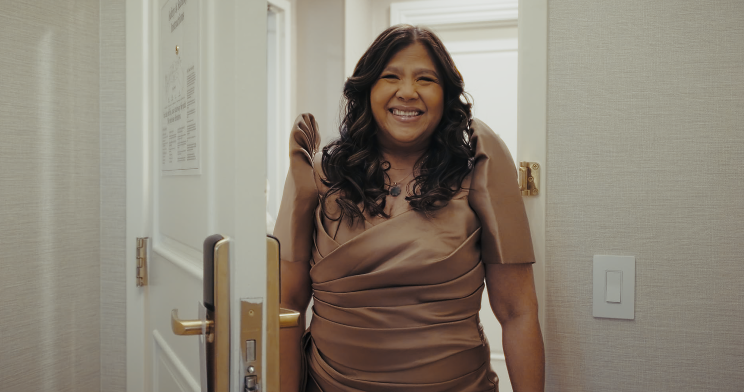 A smiling woman in a bronze satin dress stands in a bright doorway.