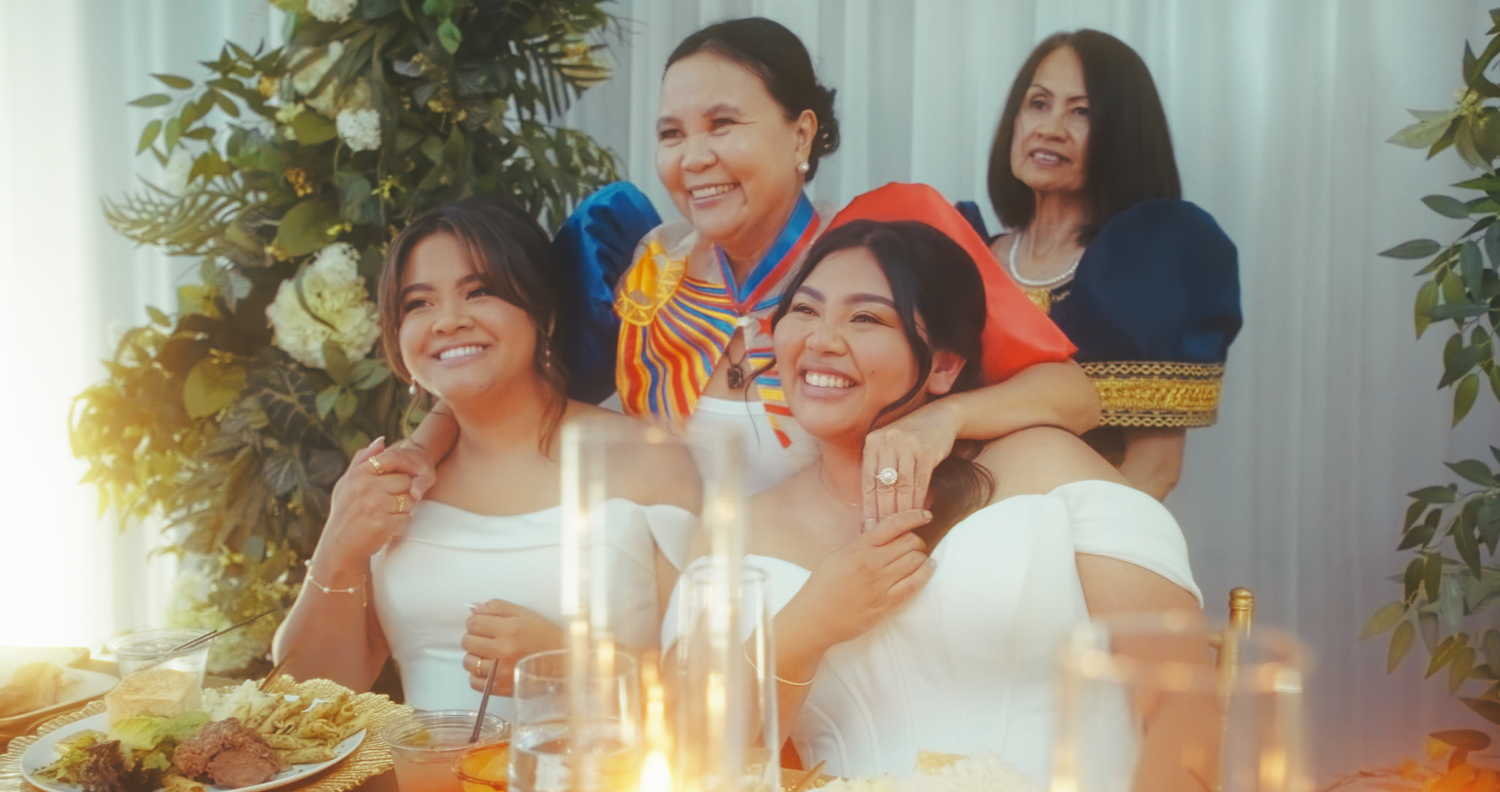 Four women in white and colorful dresses smiling together at a candlelit wedding reception table.