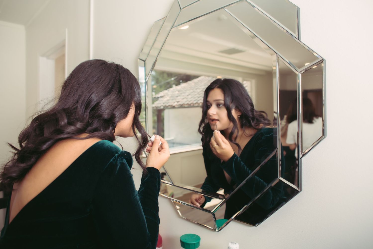 Bride applying lipstick while looking into an ornate geometric mirror during wedding prep.