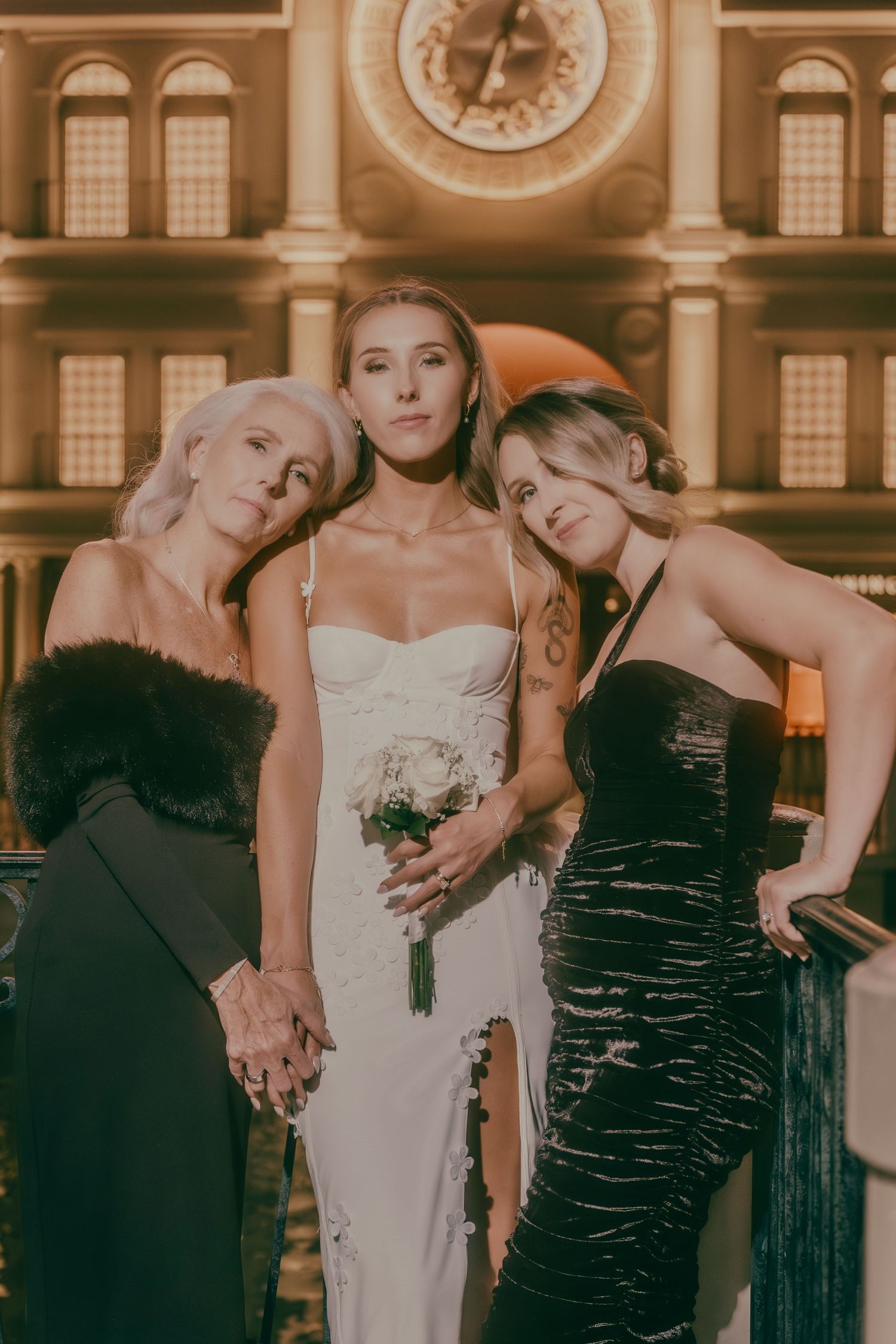 Bride with white bouquet and two women in black dresses pose before glowing clock at Las Vegas casino.