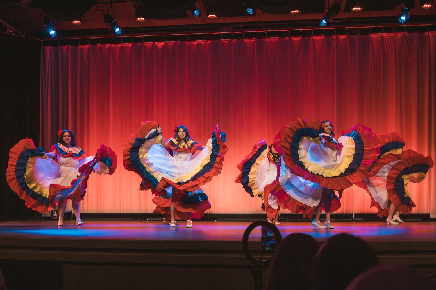 Colorful folkloric dancers in ruffled dresses perform on a red-curtained stage during a vibrant cultural dance show.
