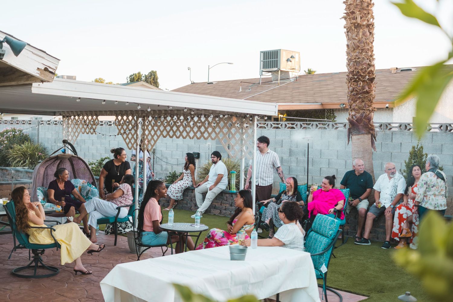 Guests socializing at an outdoor backyard gathering under a white pergola with patio furniture and palm trees.