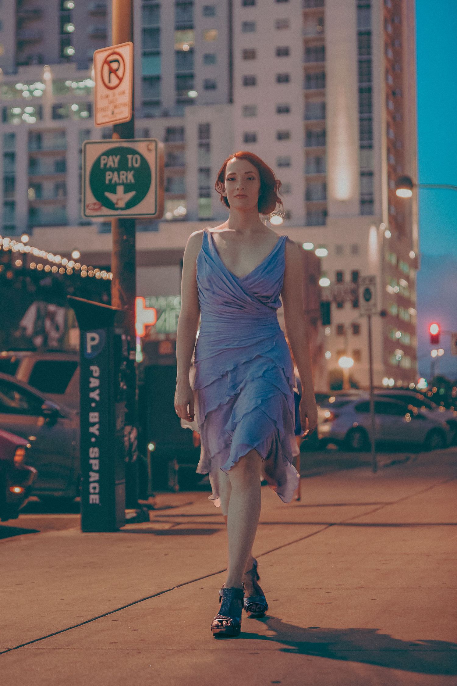 Woman in blue ruffled dress walking on urban sidewalk at night near parking signs with city buildings in background.