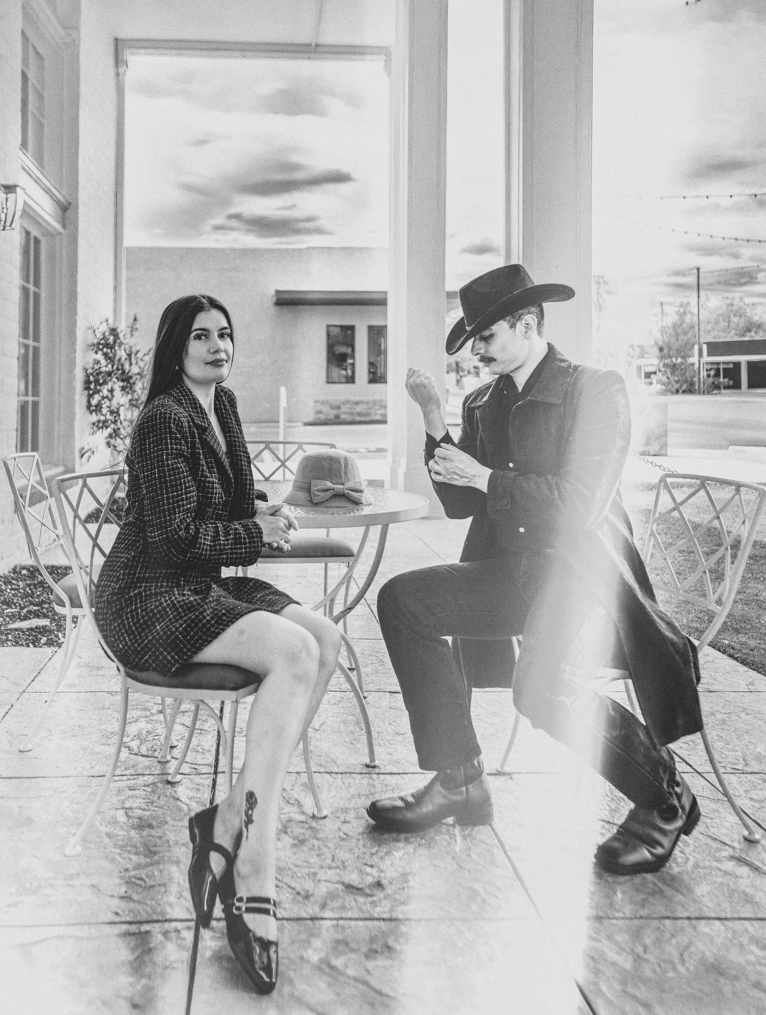 Black and white photo of a stylish couple sitting at an outdoor cafe table, dressed in vintage-inspired fashion.