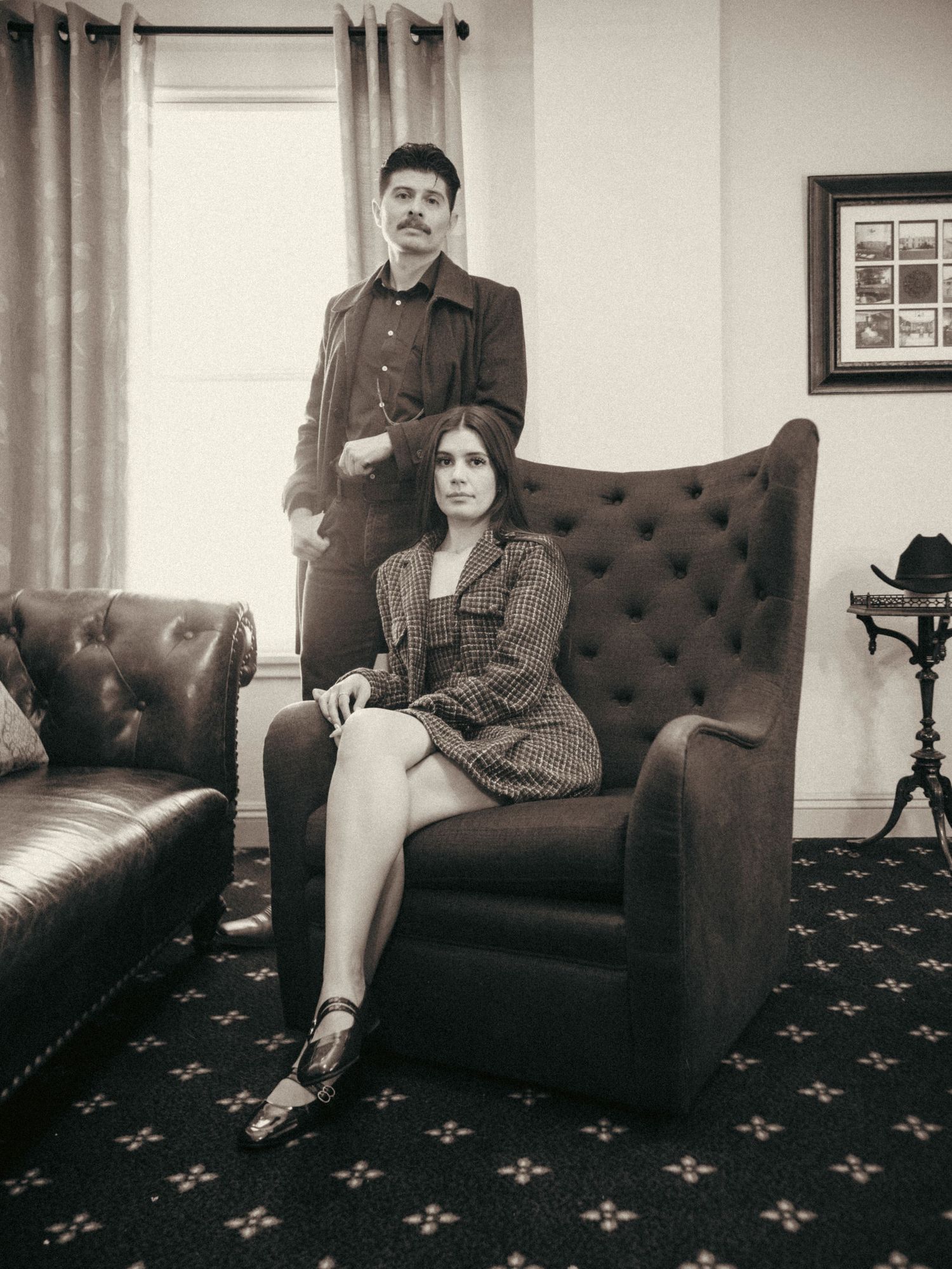 A man and woman pose in a vintage-style sepia photo, she sits in a tufted chair while he stands behind her.
