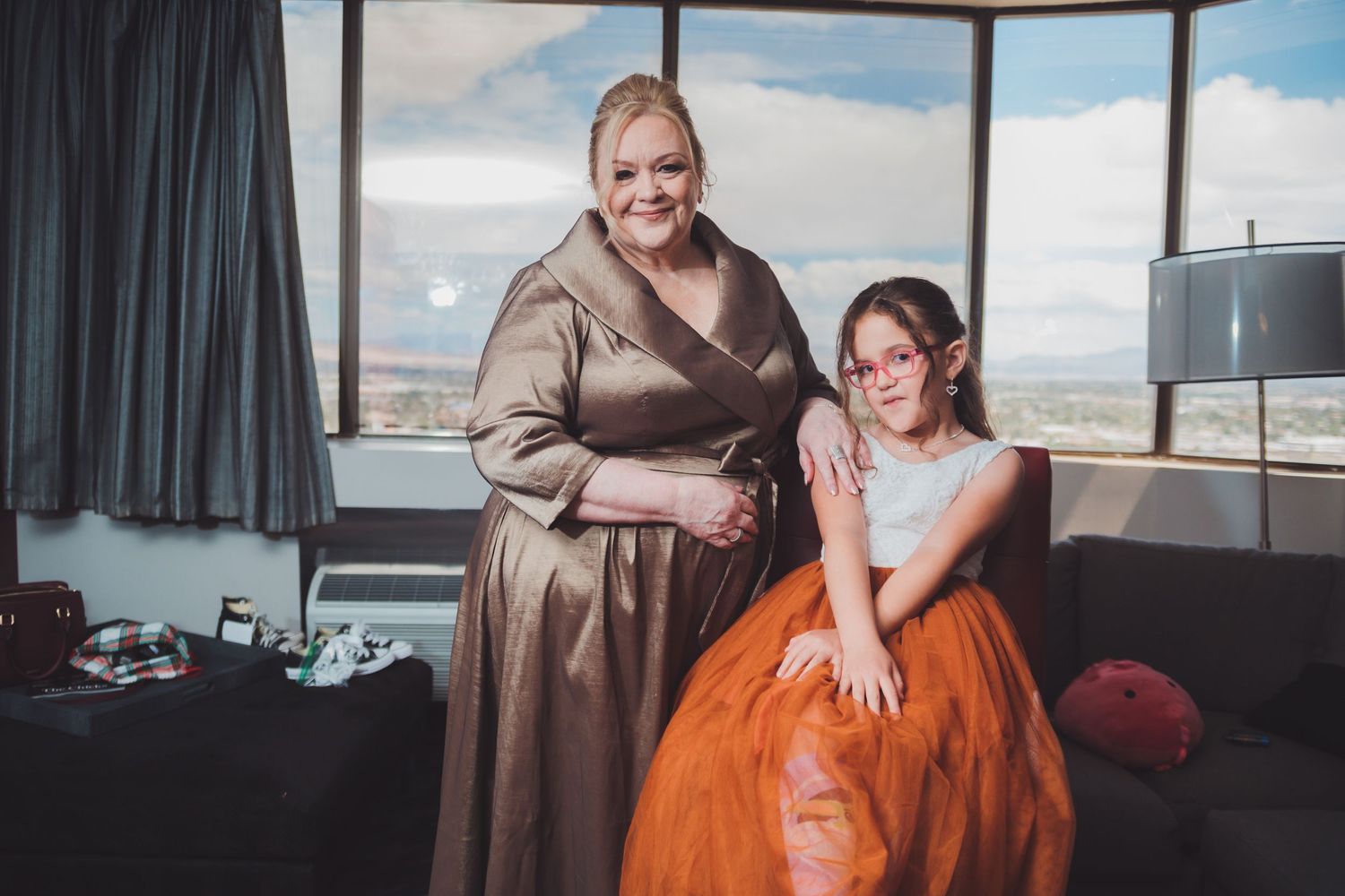 Woman in gold dress and girl in orange tulle skirt pose together in a hotel room with city views.