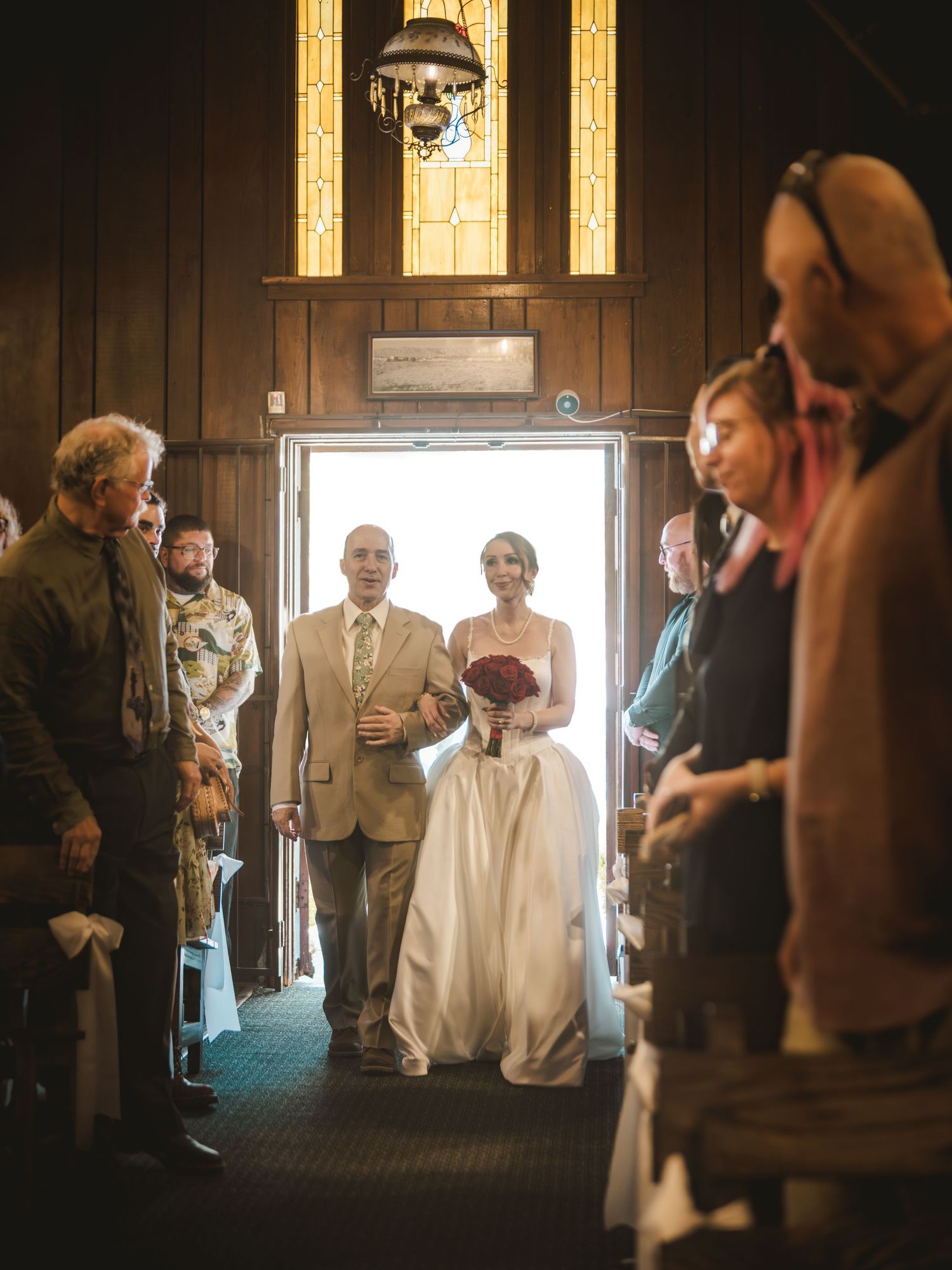 Bride in white gown holds red bouquet as she walks down the aisle with her father in a wood-paneled church.