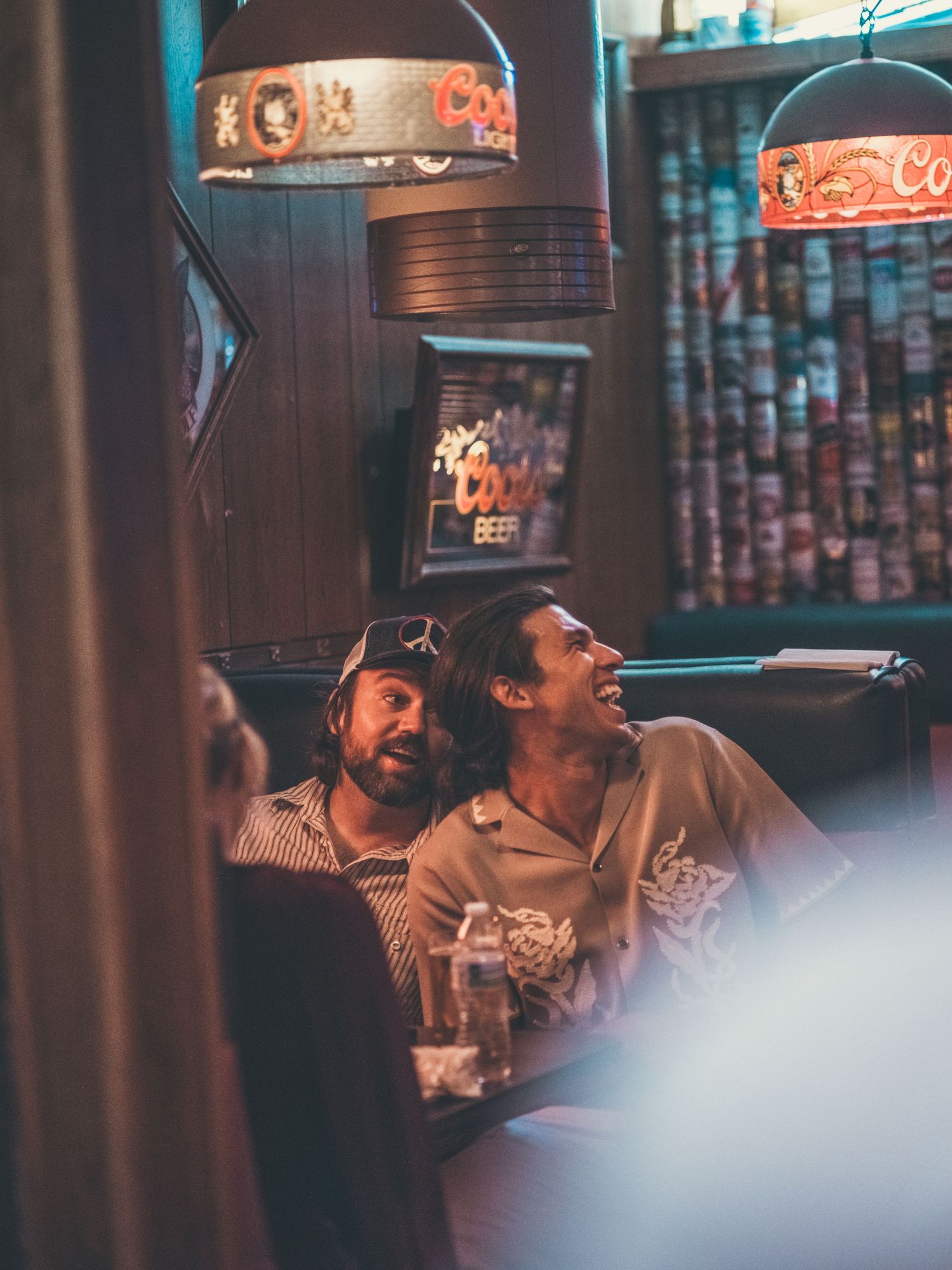 Two men laughing together in a dimly lit bar booth with vintage Coors beer signs and neon lighting.