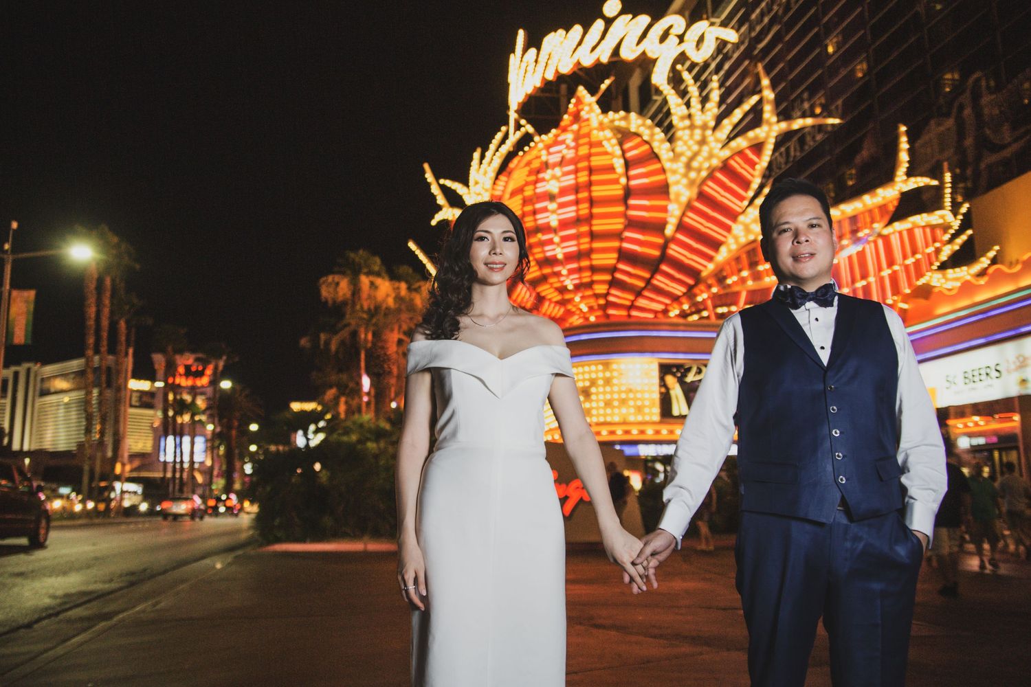 Couple in wedding attire holding hands in front of the iconic Flamingo neon sign on the Las Vegas Strip at night.