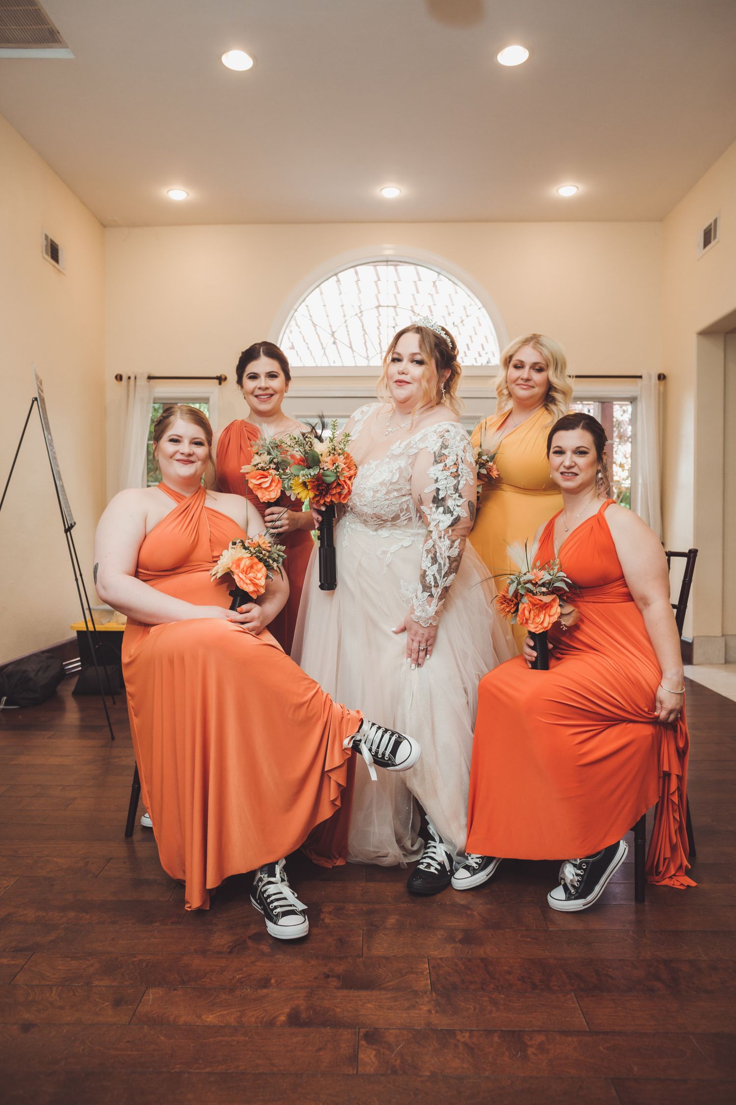 Bride with bridesmaids in orange dresses and Converse sneakers posing indoors at wedding.